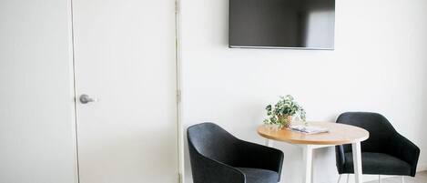 Een kamer met een witte deur, een wandtelevisie, een ronde houten tafel en twee zwarte stoelen.