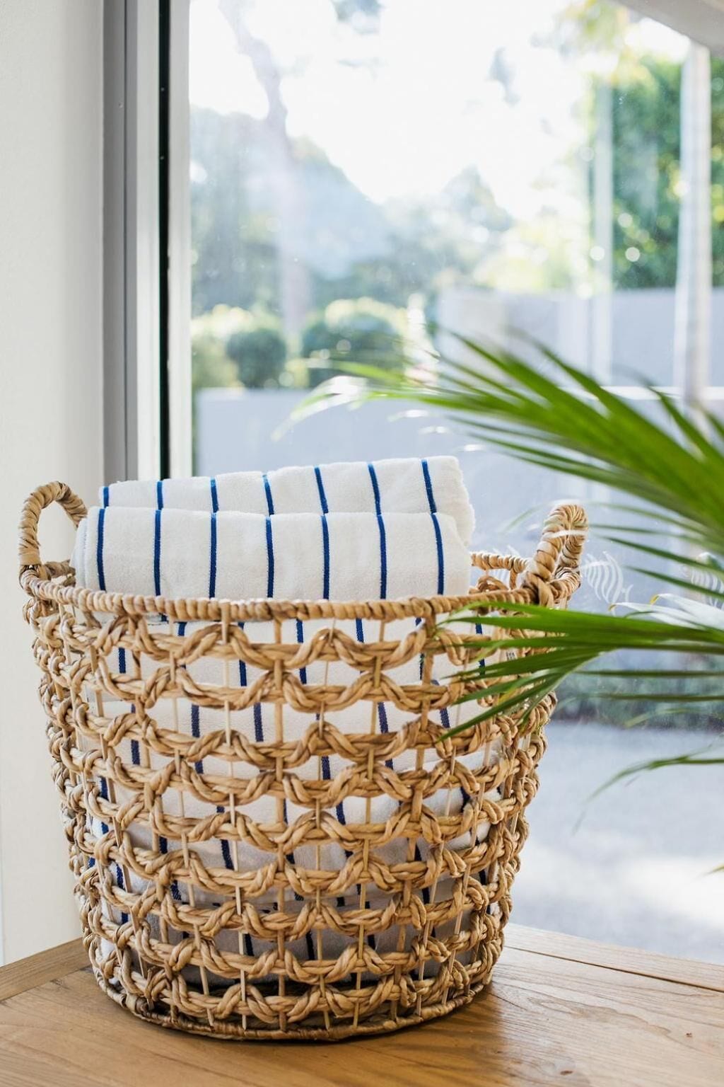 Una cesta tejida con una toalla blanca en su interior, sobre una superficie de madera cerca de una ventana con vistas a la vegetación.
