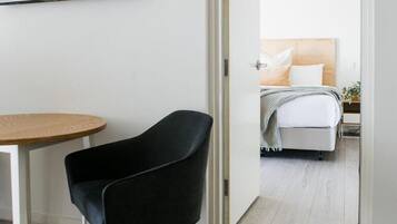 A modern hotel room with a flat-screen TV mounted on the wall, a round wooden table, a black chair, and a bed with a wooden headboard.