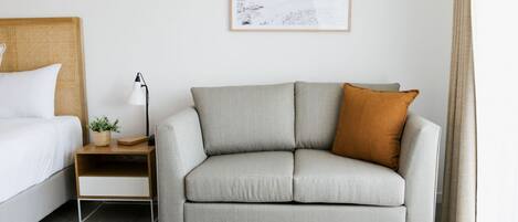 Un dormitorio moderno con un sofá gris, una mesita de noche con lámpara, un cuadro enmarcado en la pared y una cama con ropa de cama blanca.