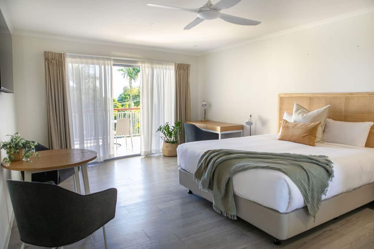 A hotel room with a bed, a desk, a chair, a ceiling fan, and a view of the outdoors.