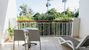 Enjoy a tranquil retreat with a balcony overlooking lush greenery and palm trees, featuring a table for two and a comfortable lounge chair.
