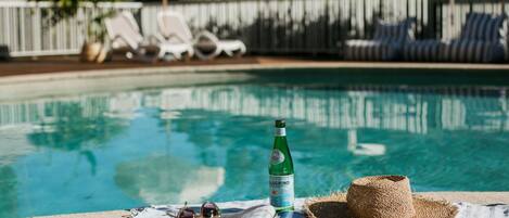 A poolside area with lounge chairs, a bottle of alcohol, sunglasses, and a hat on a towel.