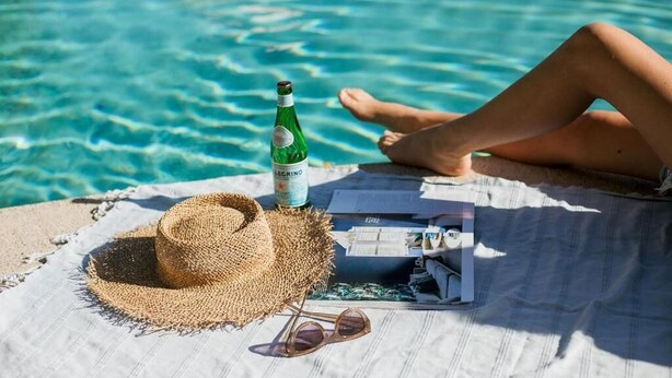 Una persona relaxant-se al costat d'una piscina amb un barret de palla, ulleres de sol, una ampolla i una revista sobre una tovallola.