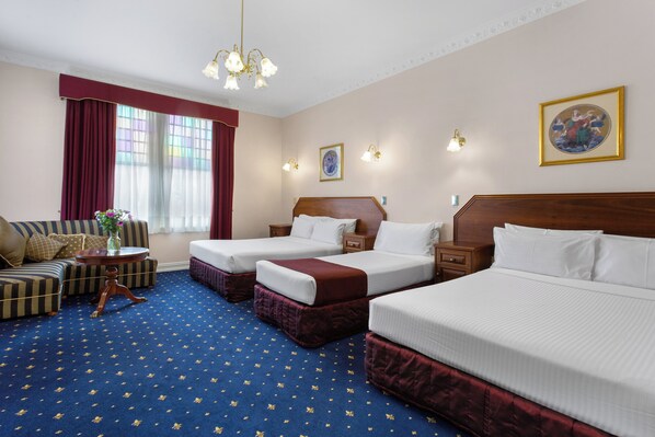 Habitación de hotel con dos camas individuales, un sofá, una mesita con un florero y un amplio ventanal con cortinas.