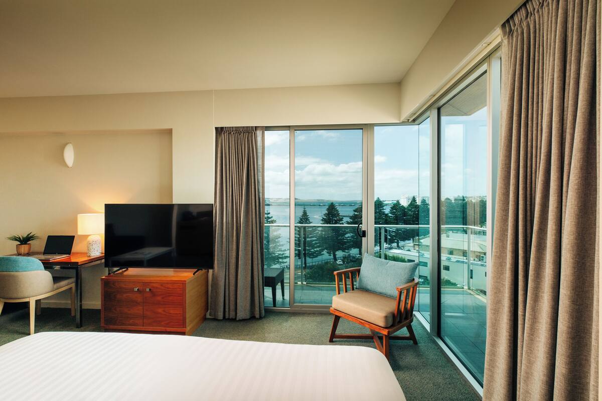 Ein Hotelzimmer mit Bett, Schreibtisch mit Laptop, Fernseher, Stuhl und einem Balkon mit Stadtblick.