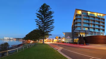 A modern hotel with a large tree in front, a waterfront view, and a well-lit entrance.