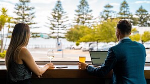 Duas pessoas sentadas à mesa junto a uma janela, com um laptop e bebidas, com vista para uma rua com carros e árvores.