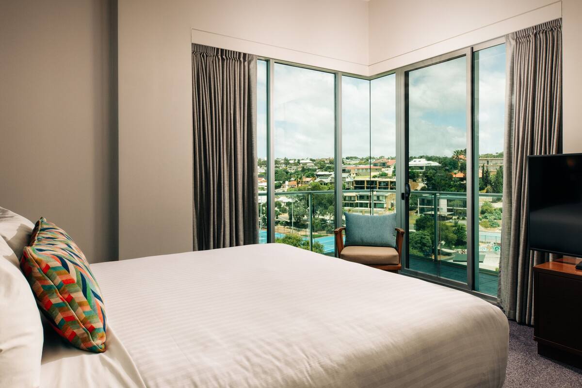 Ein Hotelzimmer mit einem großen Bett, einem Sessel, einem Fernseher und Blick auf eine Stadtlandschaft durch große Fenster.