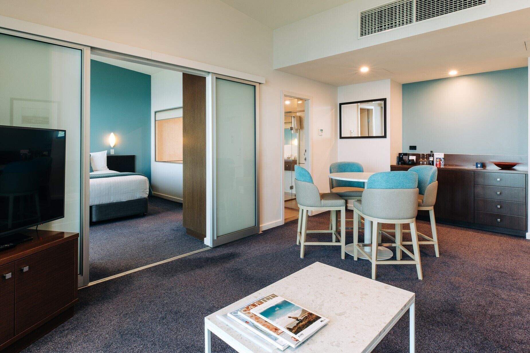 A modern hotel room with a bed, a TV, a dining area with chairs, and a kitchen counter.