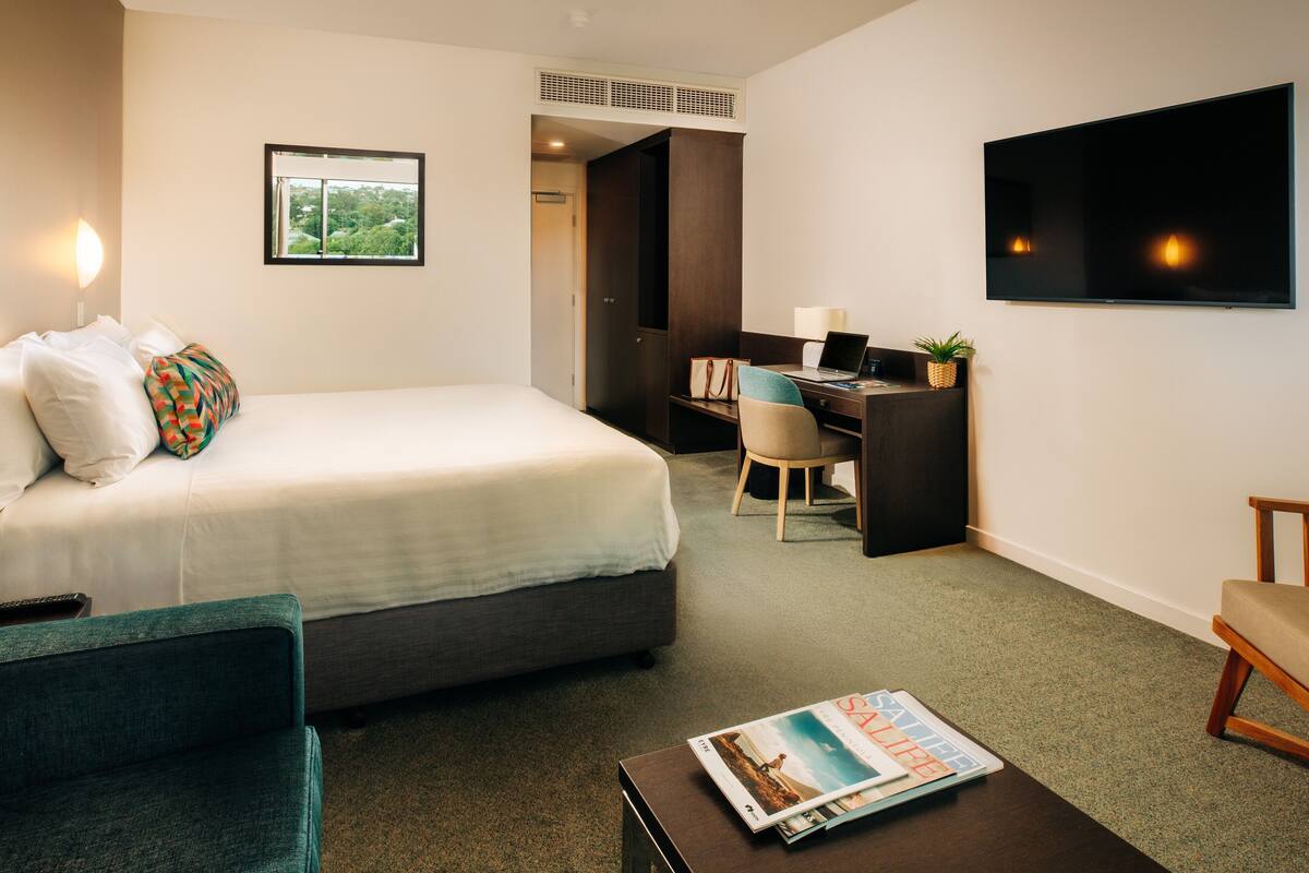 Ein Hotelzimmer mit Bett, Schreibtisch, Stuhl, Fernseher und einer Zeitschrift auf dem Couchtisch.