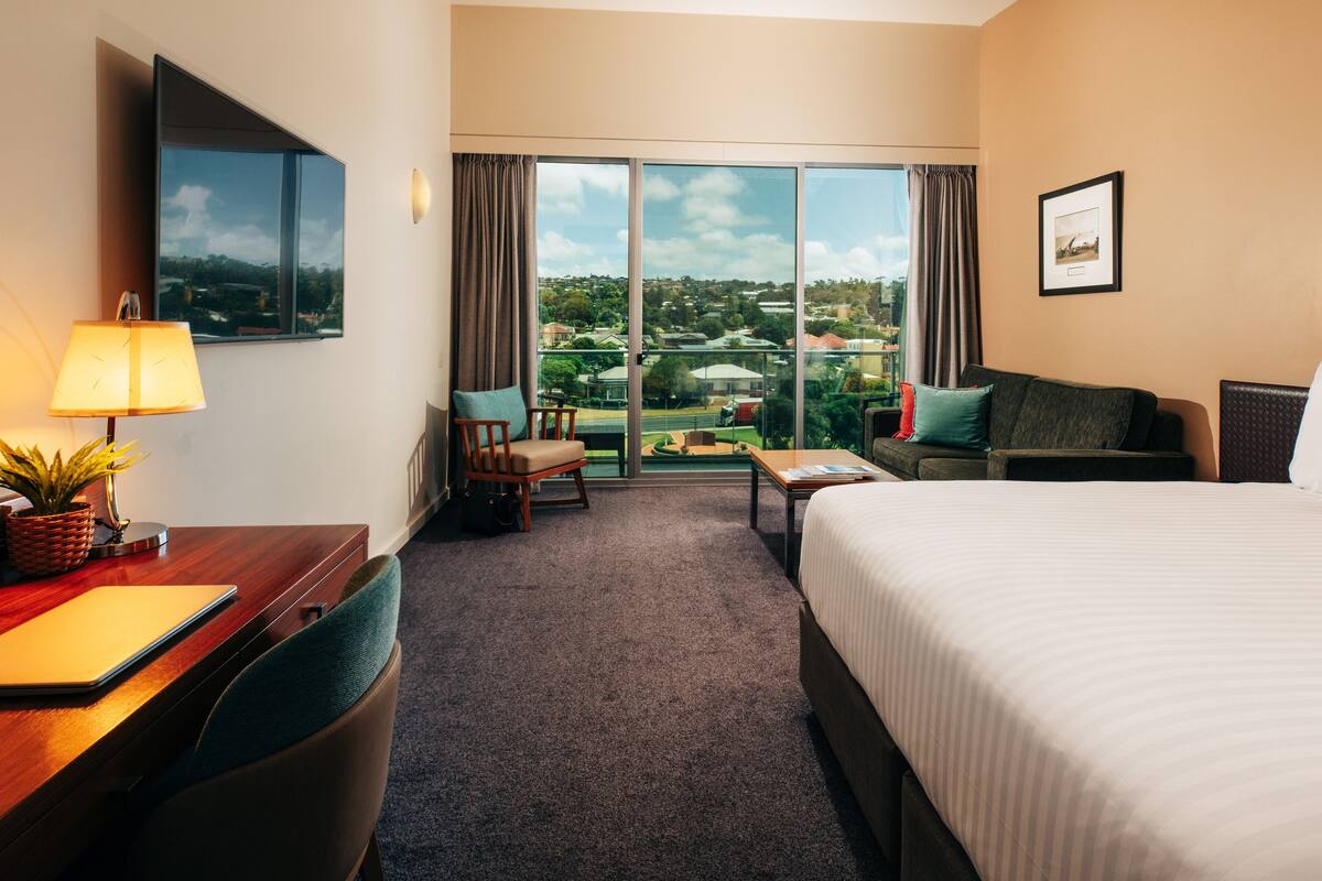 Ein Hotelzimmer mit Bett, Schreibtisch, Stuhl und Blick auf die Stadt durch große Fenster.