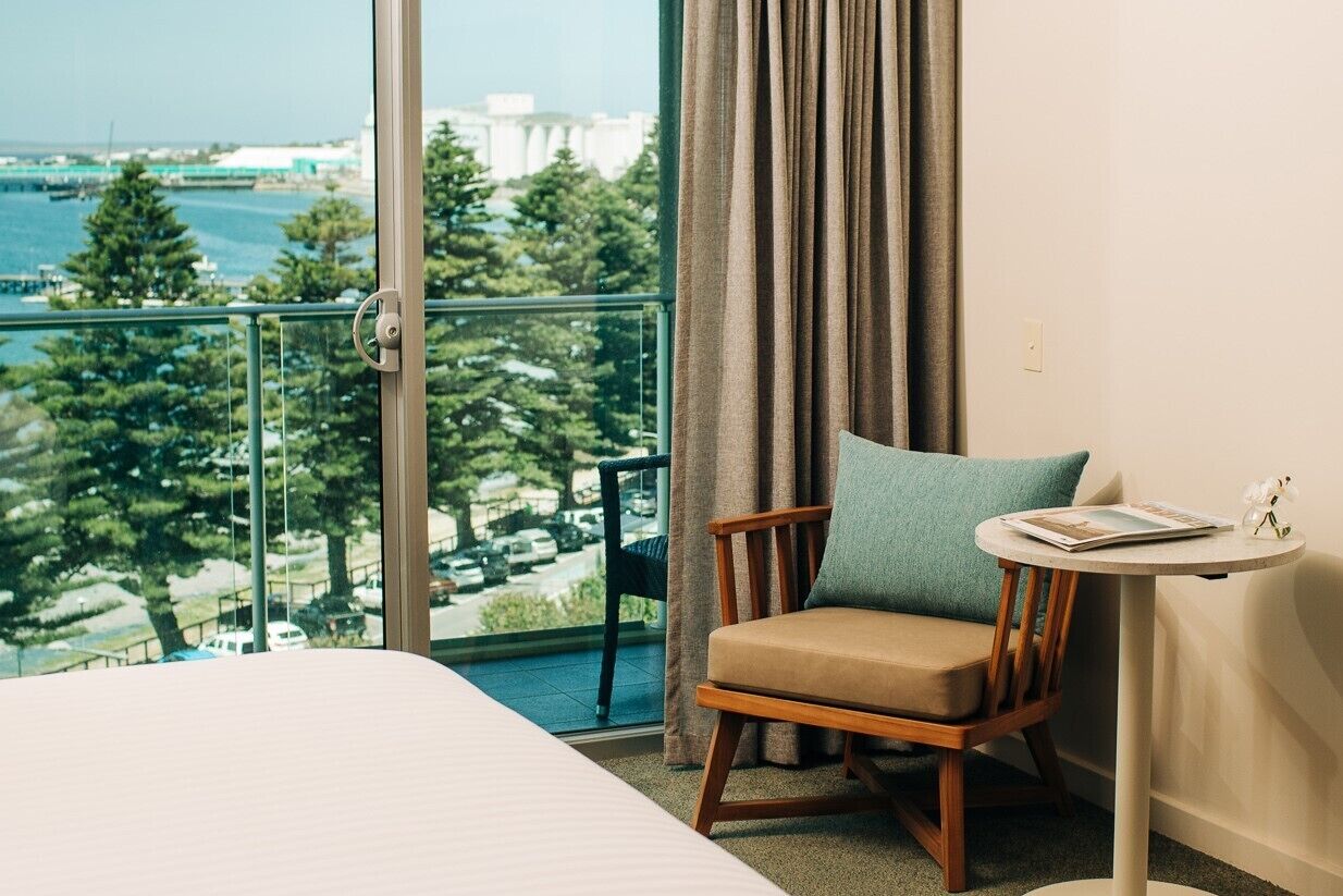 A hotel room with a bed, a chair, a small table, and a view of the sea and buildings.