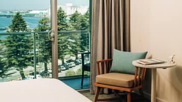 A hotel room with a bed, a chair, a small table, and a view of the sea and buildings.