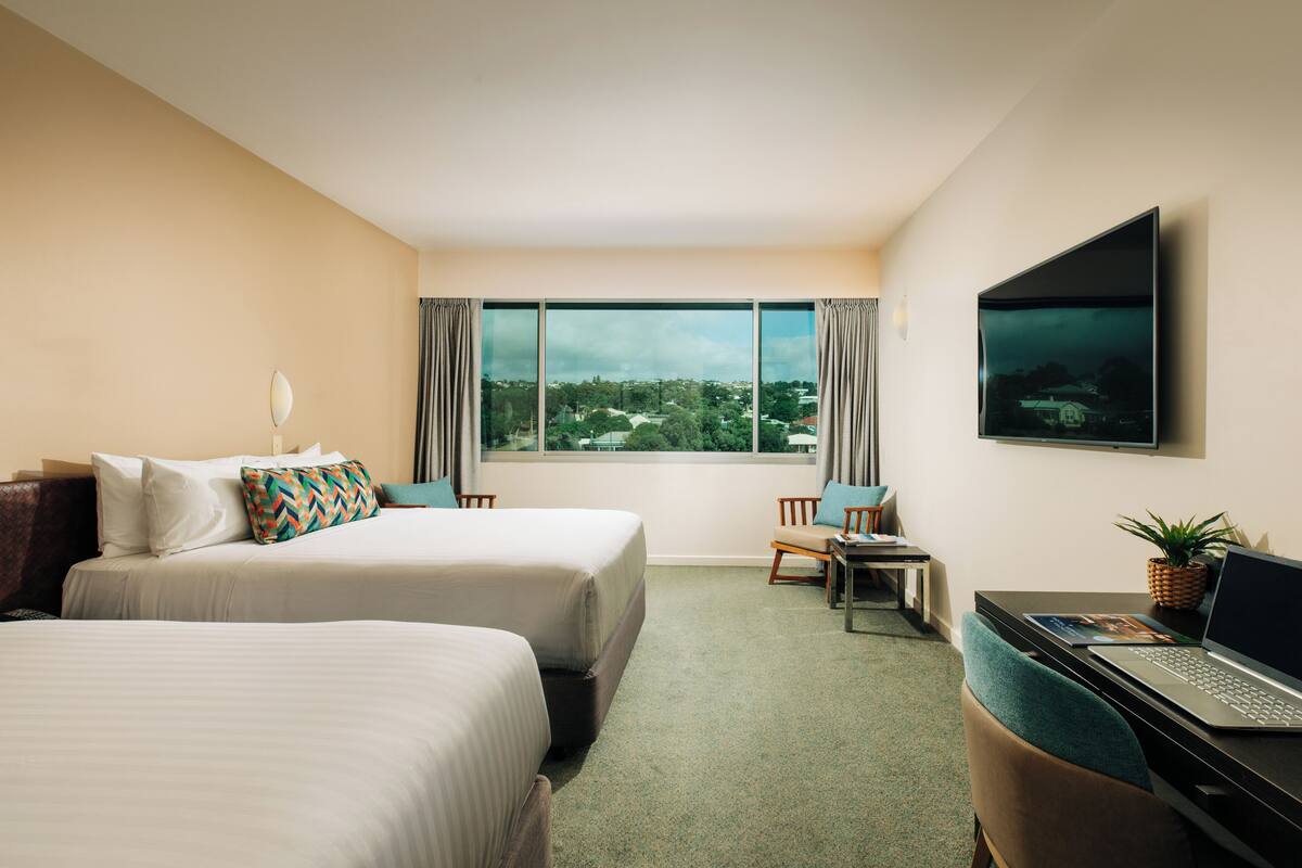 Ein Hotelzimmer mit zwei Betten, einem Schreibtisch mit einem Laptop, einem Fernseher und einem großen Fenster mit Blick auf die Stadt.