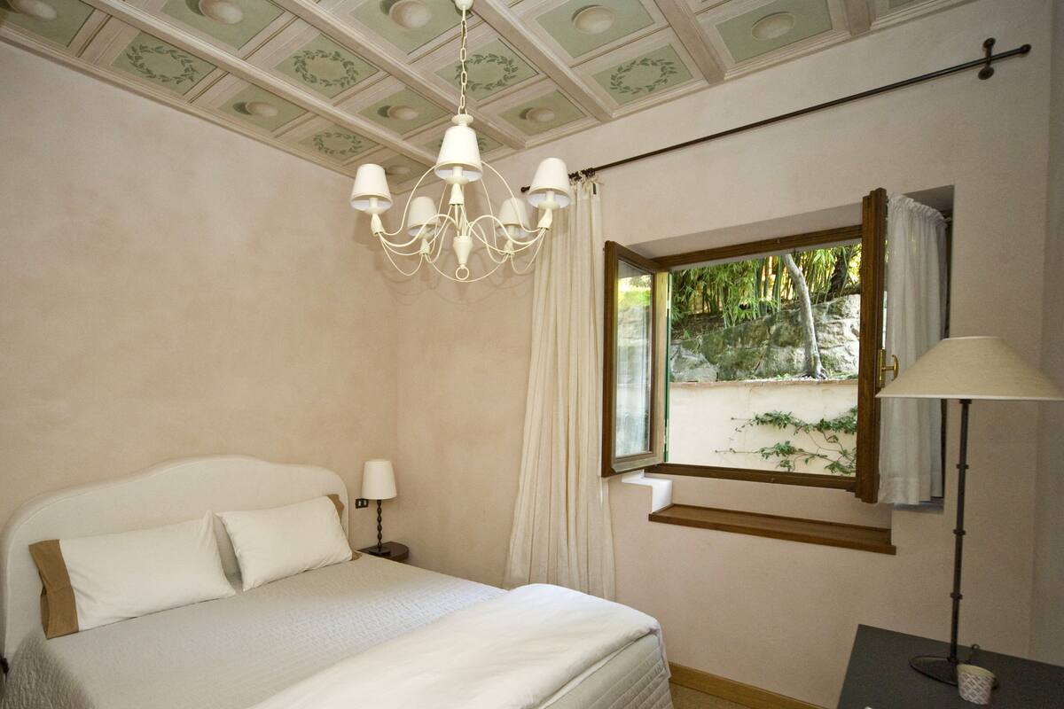 Un dormitorio con cama, candelabro, ventana con cortinas y mesita de noche.