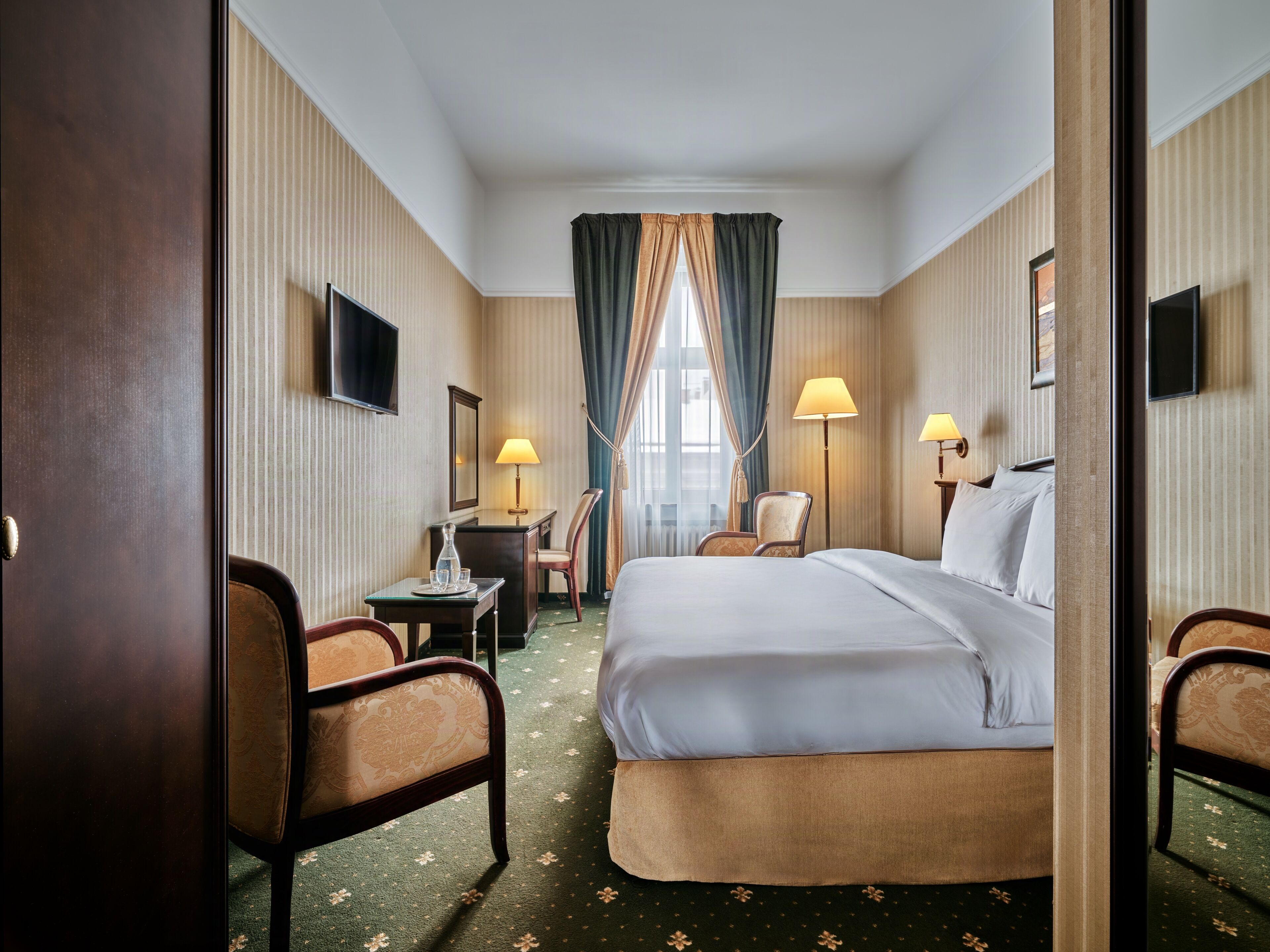 A hotel room with a bed, two chairs, a desk, a TV, and a window with curtains.