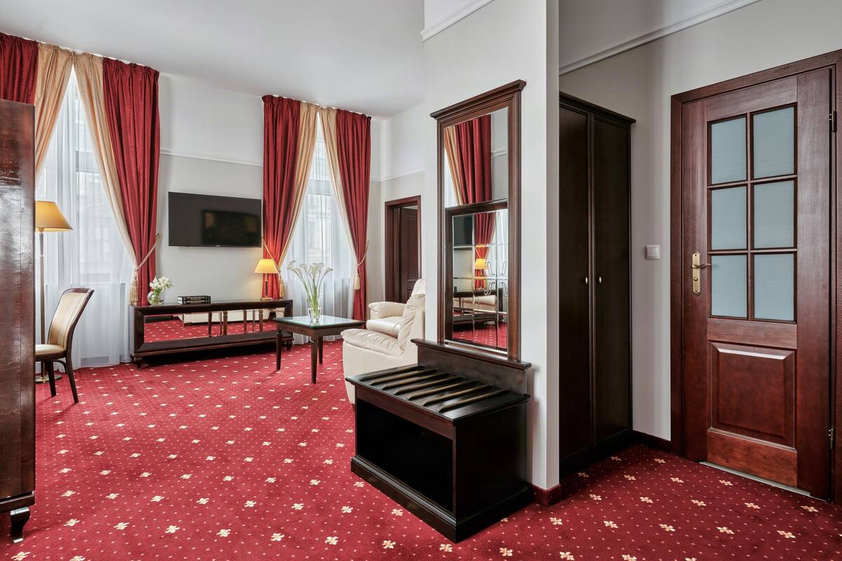 Una habitación de hotel con alfombra roja, un sofá, una mesa con lámpara, un televisor y un espejo.