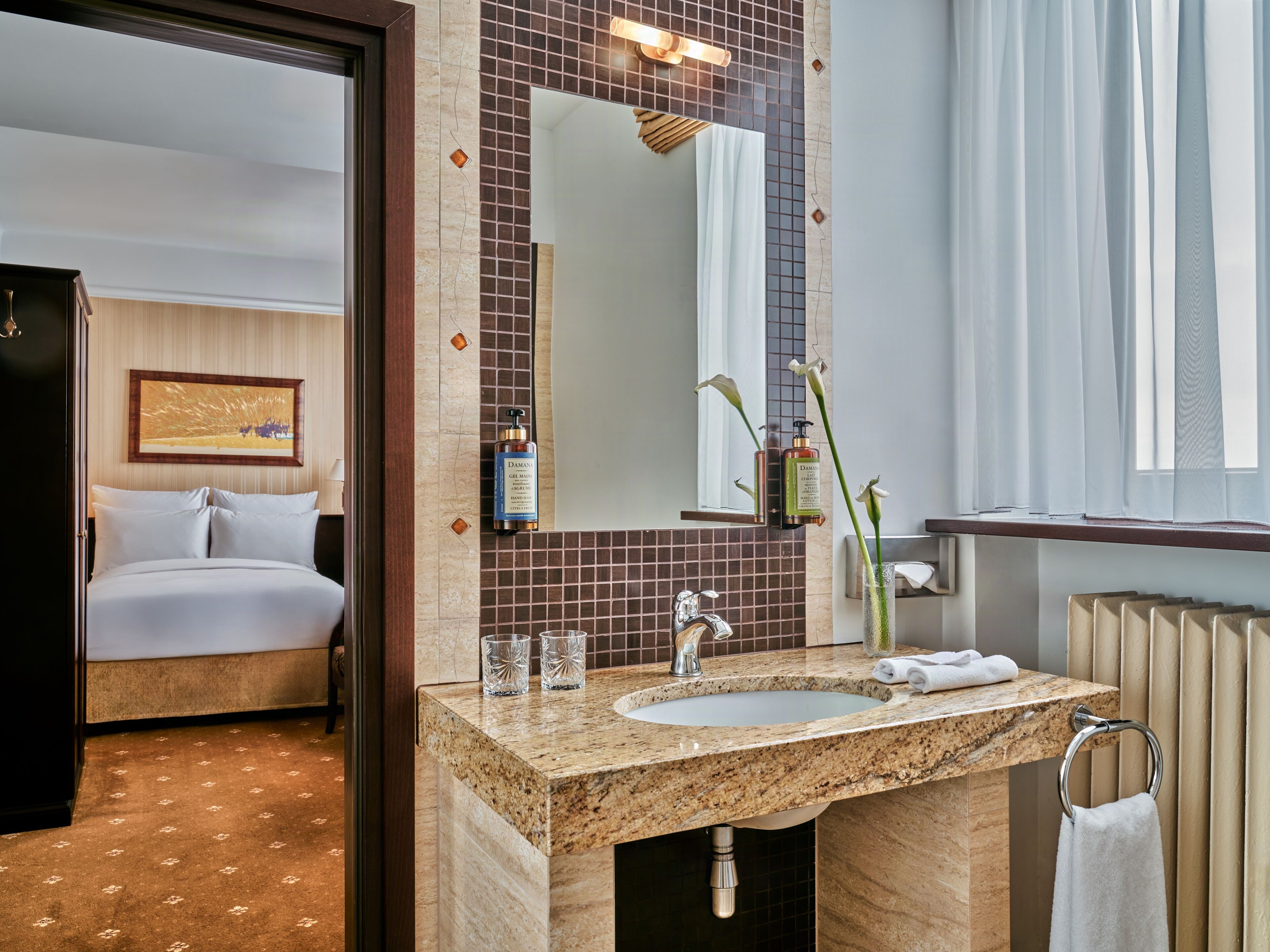 A hotel bathroom with a marble sink, a large mirror, and a bed visible through an open door.