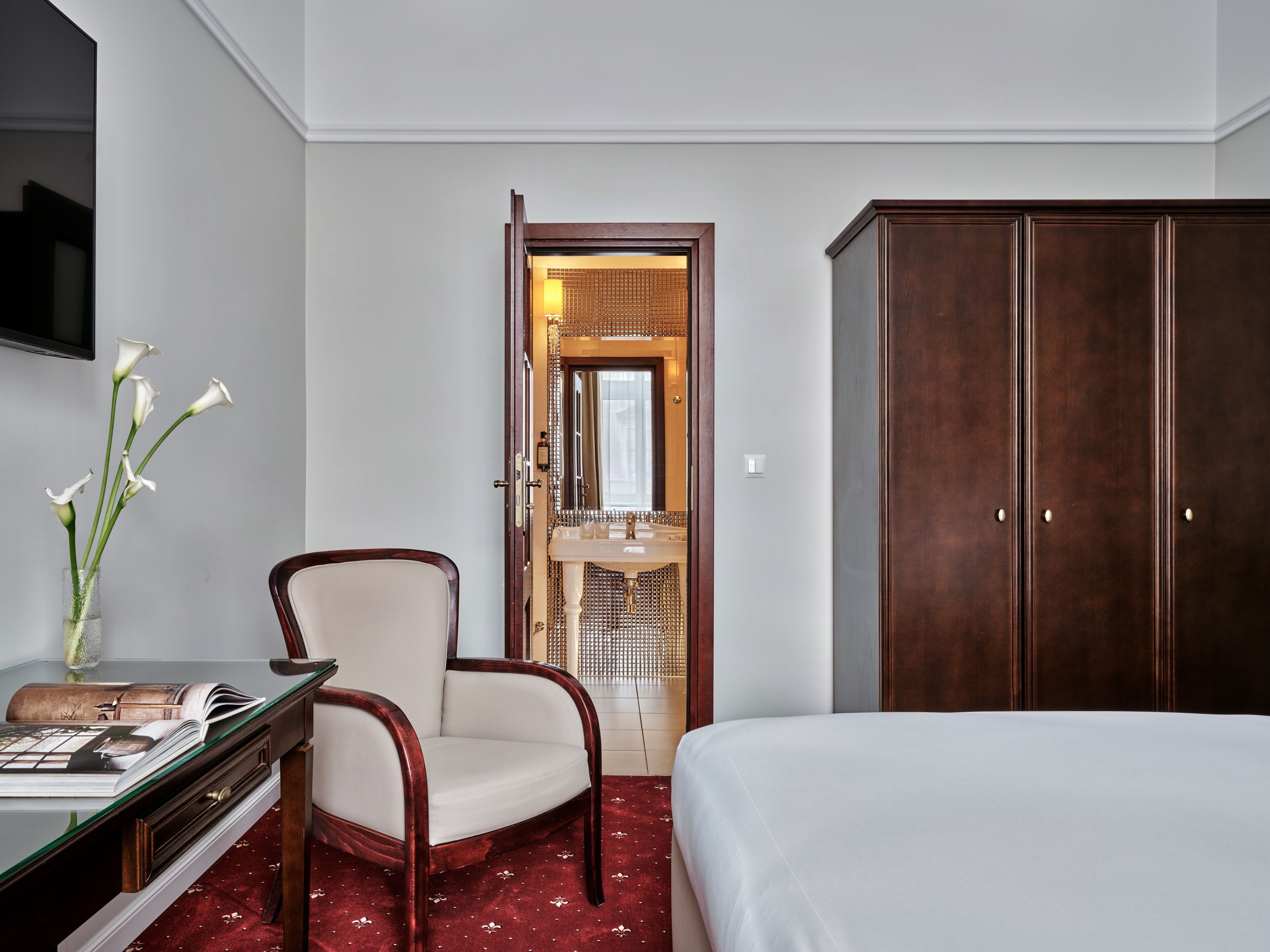 Una camera d'albergo con un letto, una scrivania con un vaso di fiori, una sedia, un armadio e un bagno visibile attraverso una porta aperta.