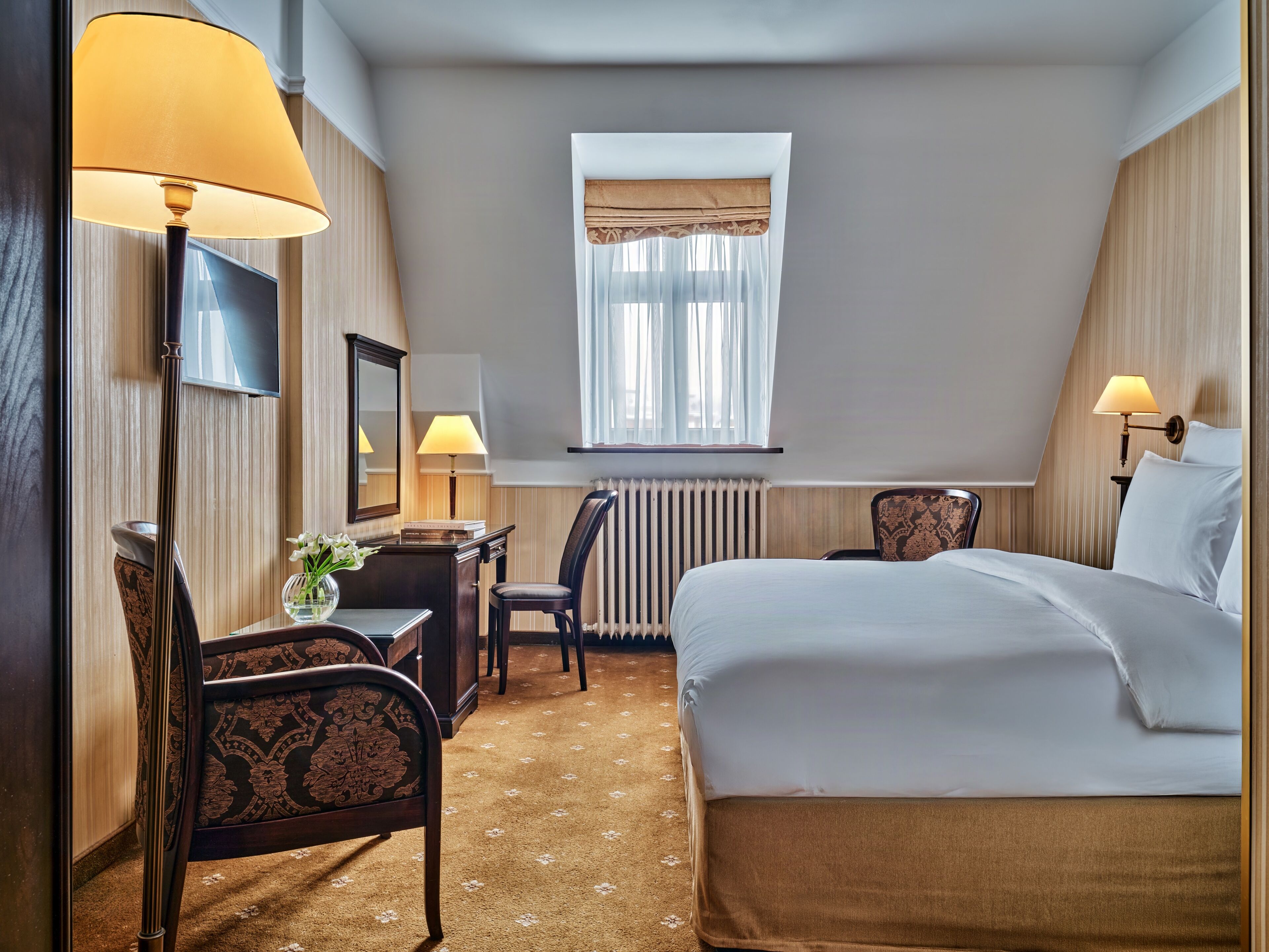 Una habitación de hotel con cama, escritorio, silla, lámpara y ventana con cortinas.