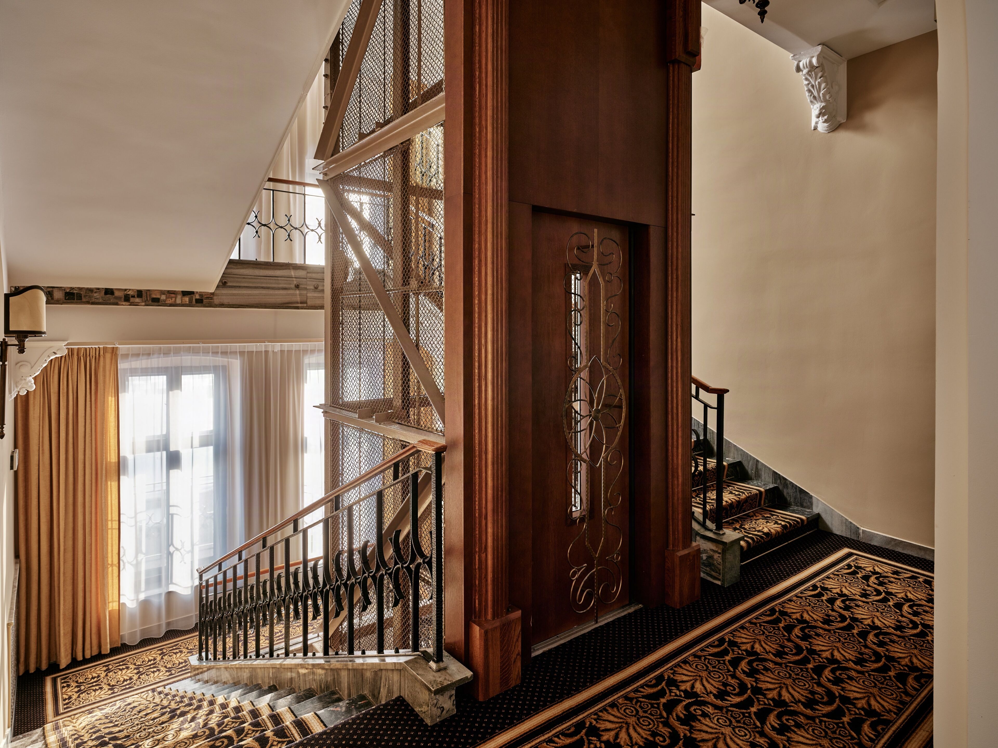 Una imponente escalera con barandales de metal ornamentados, un suelo con motivos y una puerta de madera con un panel decorativo.