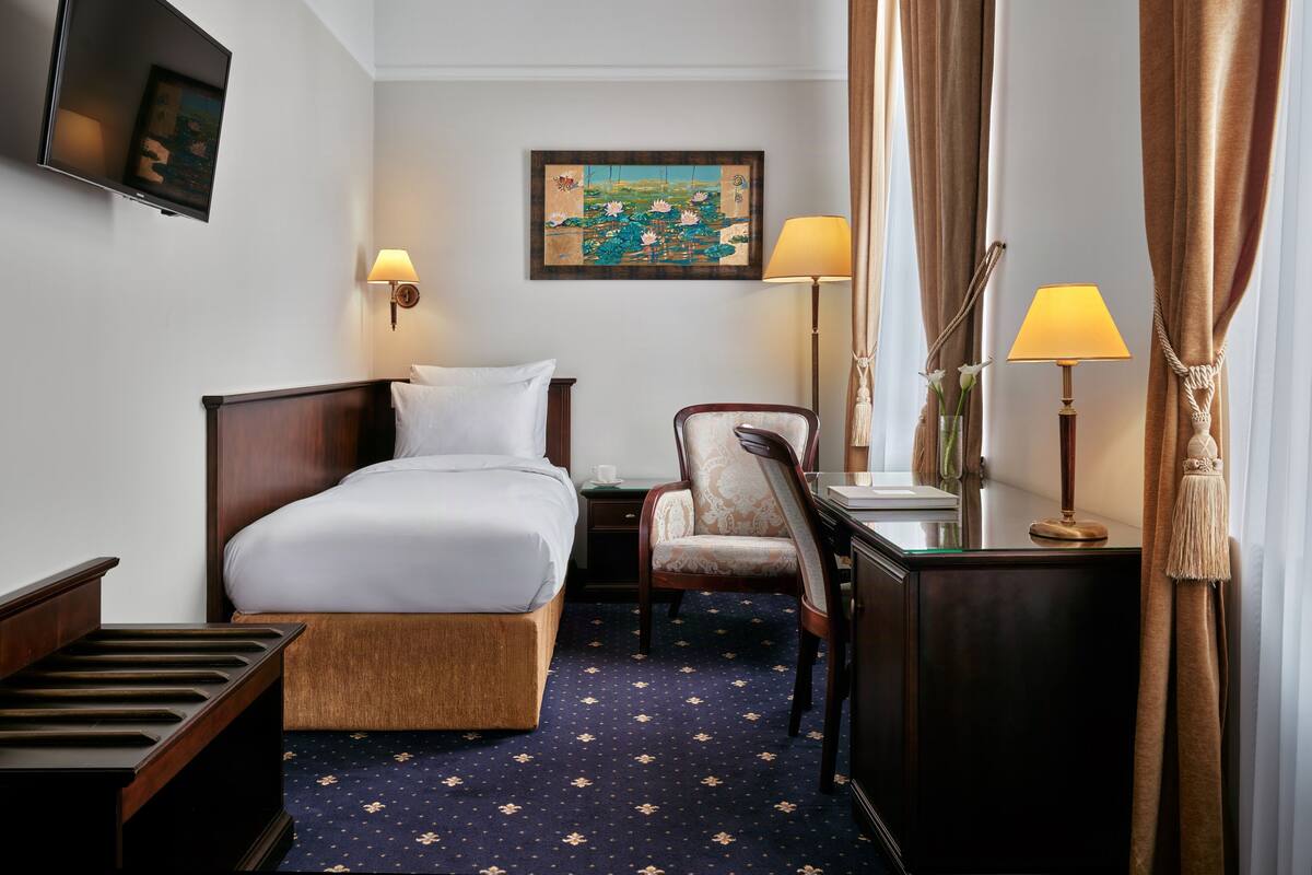 Una habitación de hotel con cama, sillón, escritorio, lámpara, un cuadro y televisión.
