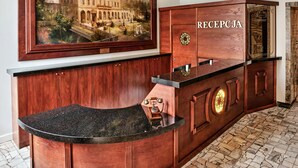 A reception desk with a 'RECEPCJA' sign, a framed painting of a building, and a chandelier. - Hotel President (Bielsko-Biala)