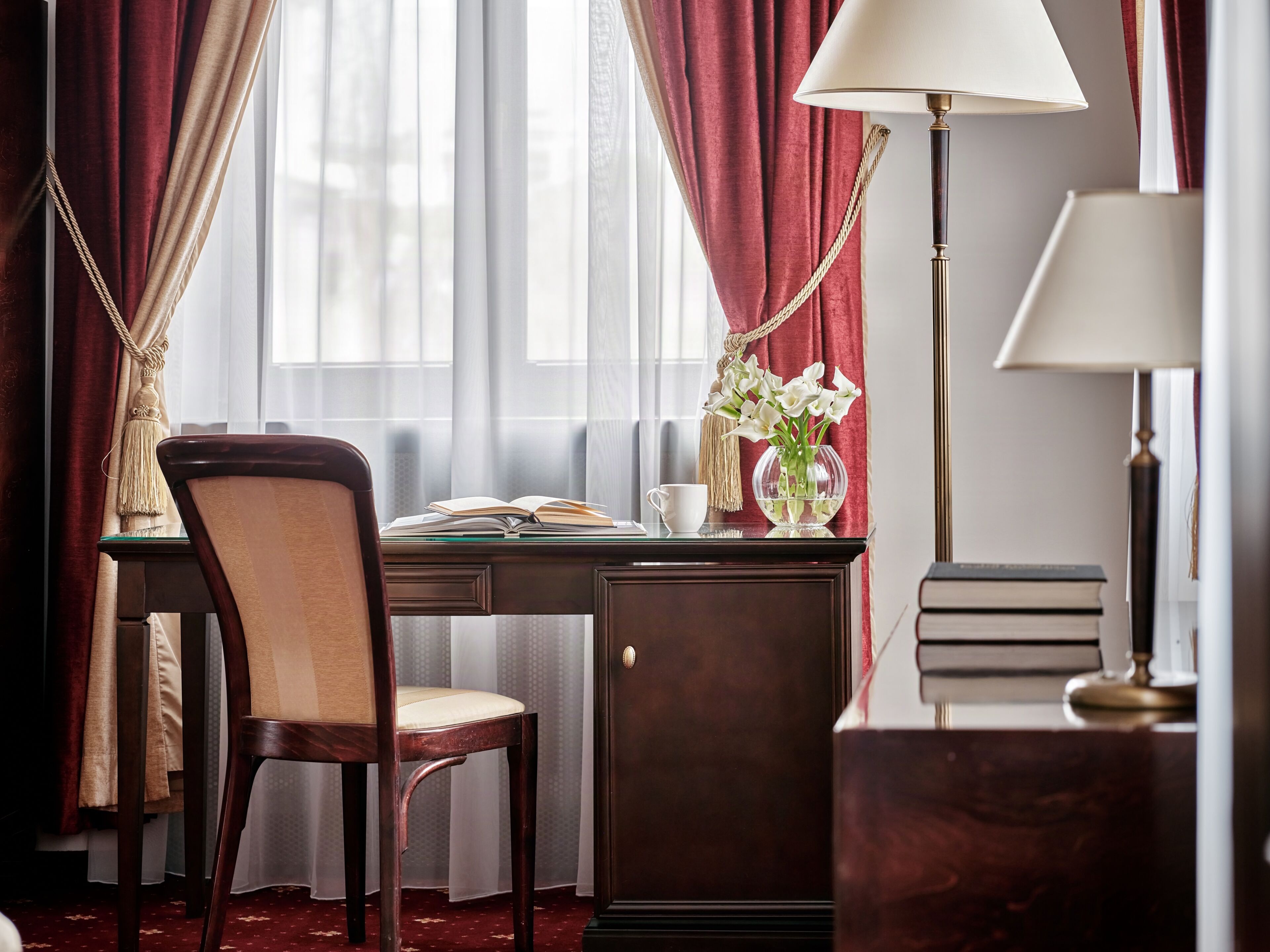 Una habitación con escritorio, silla, lámpara y ventana con cortinas.