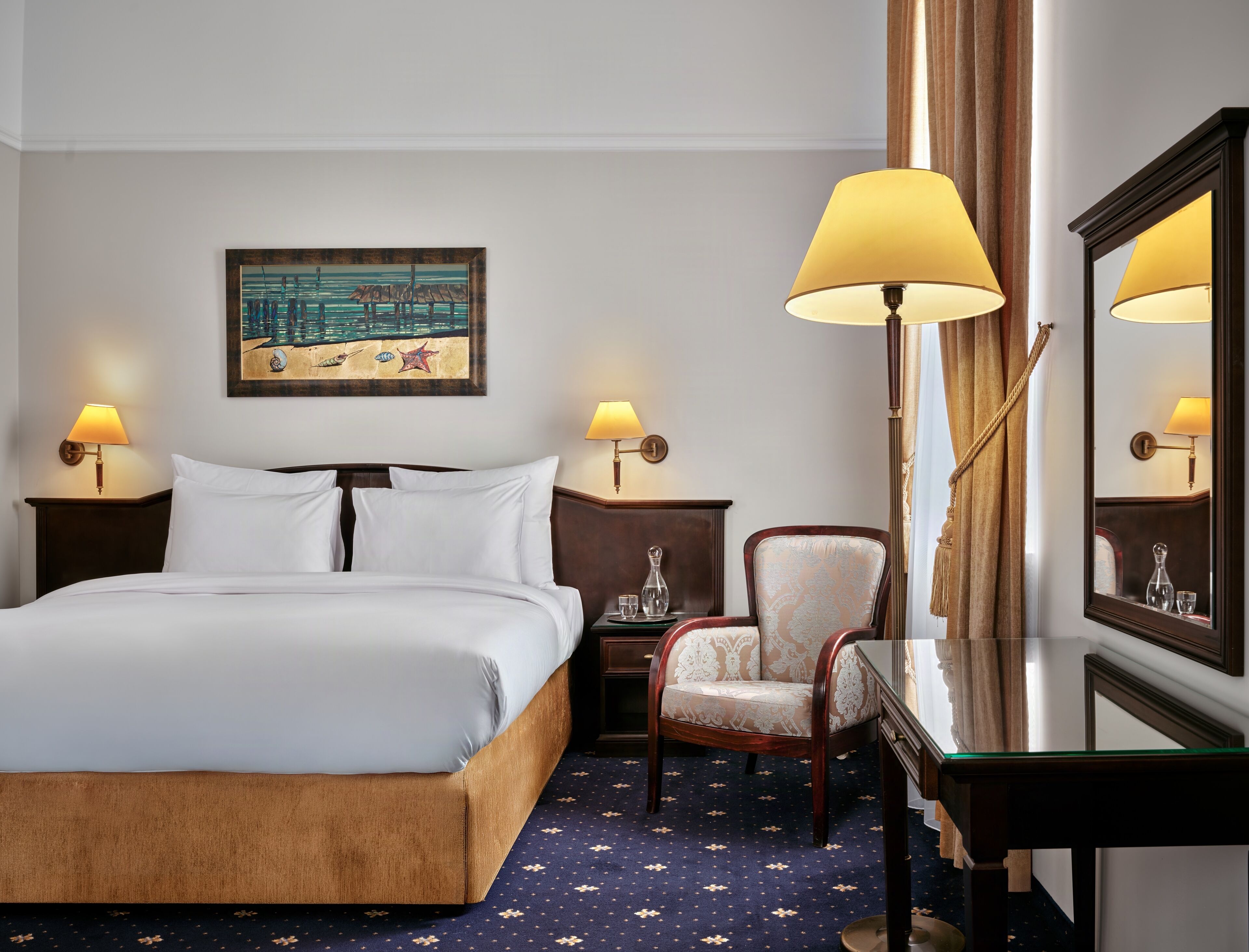A hotel room with a bed, two bedside lamps, a chair, a mirror, and a painting on the wall.