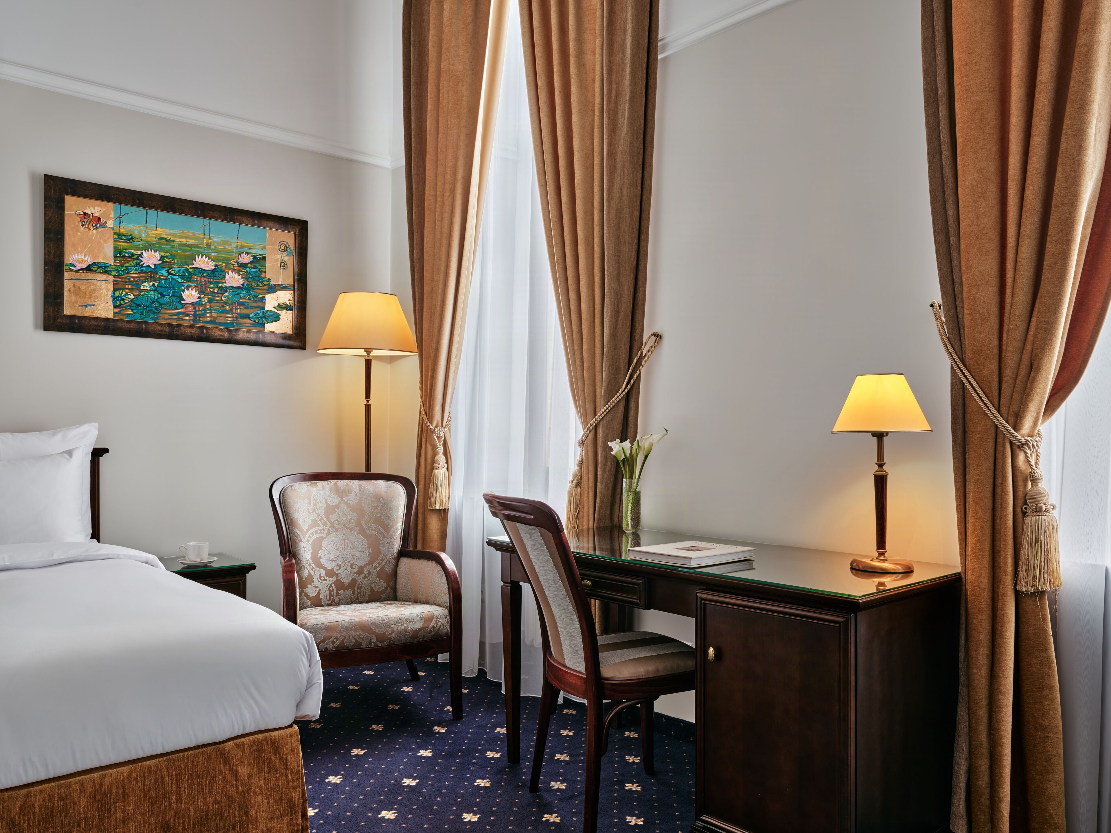 A hotel room with a bed, two chairs, a desk, a lamp, a painting, and curtains.
