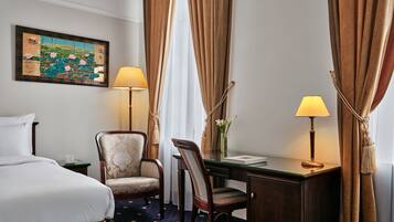 A hotel room with a bed, two chairs, a desk, a lamp, a painting, and curtains.
