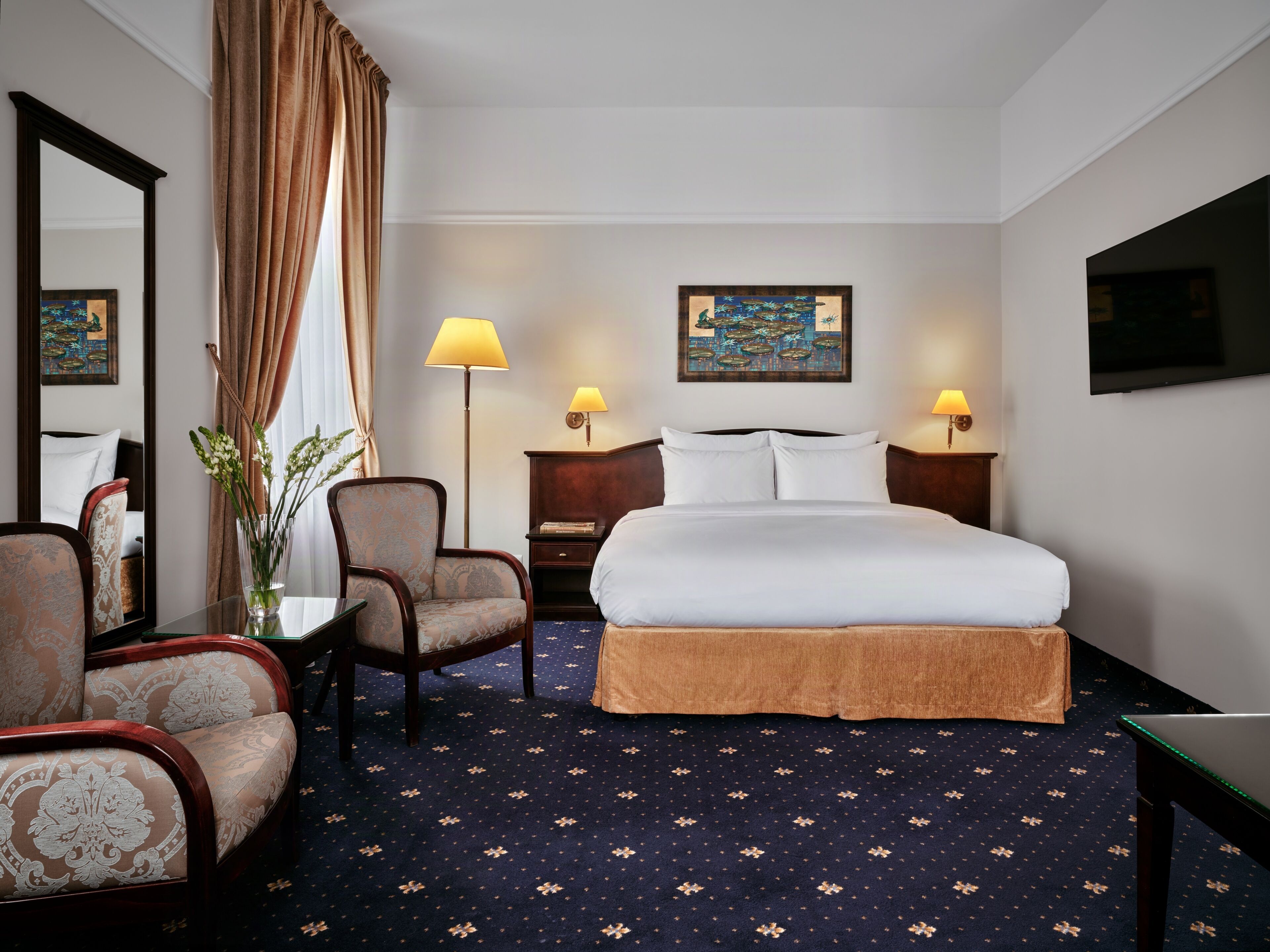 A hotel room with a bed, two chairs, a desk, a lamp, a painting, and a television.