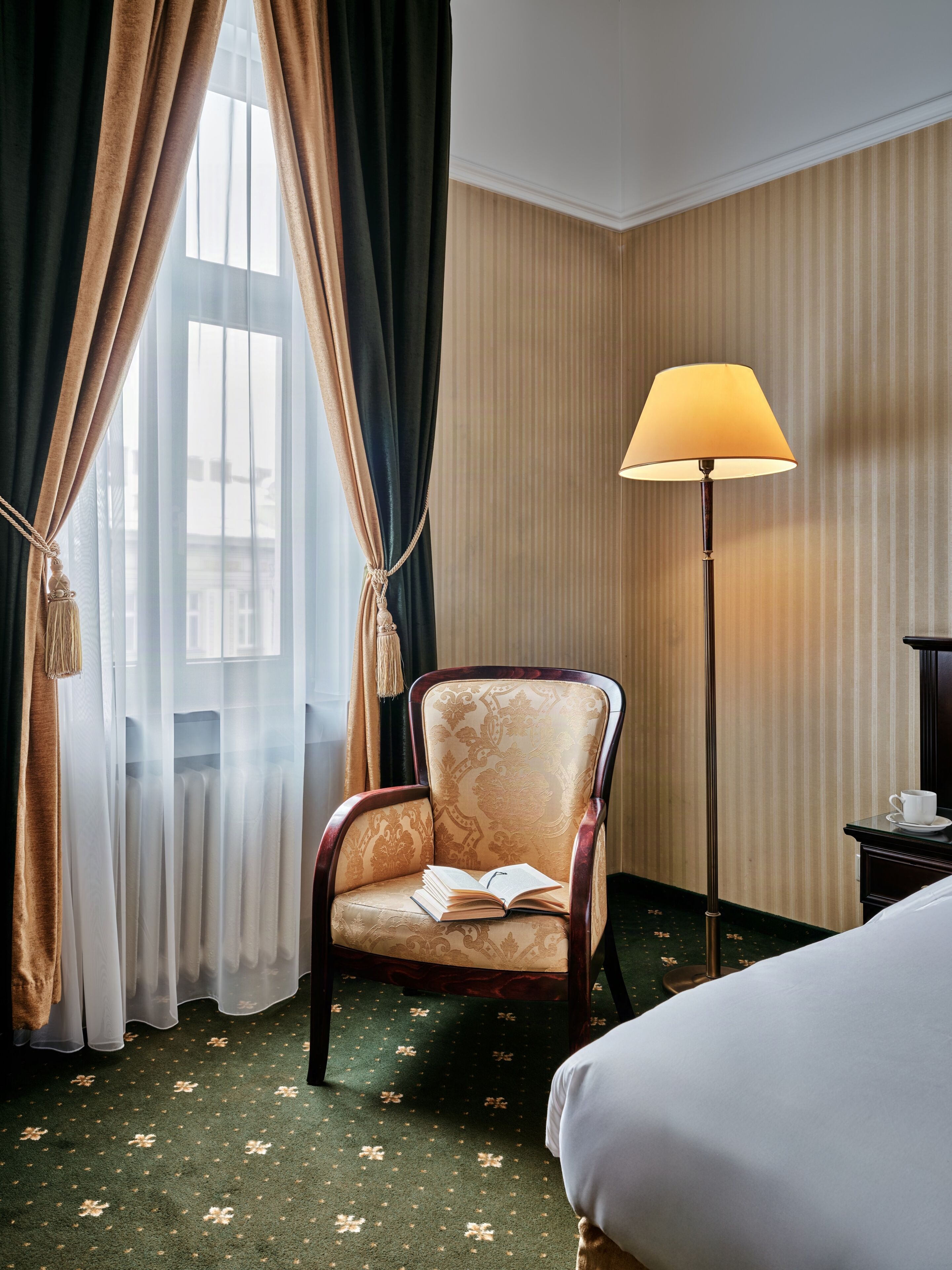 Habitación de hotel con cama, una silla con un libro abierto, una lámpara de pie y una ventana con cortinas.