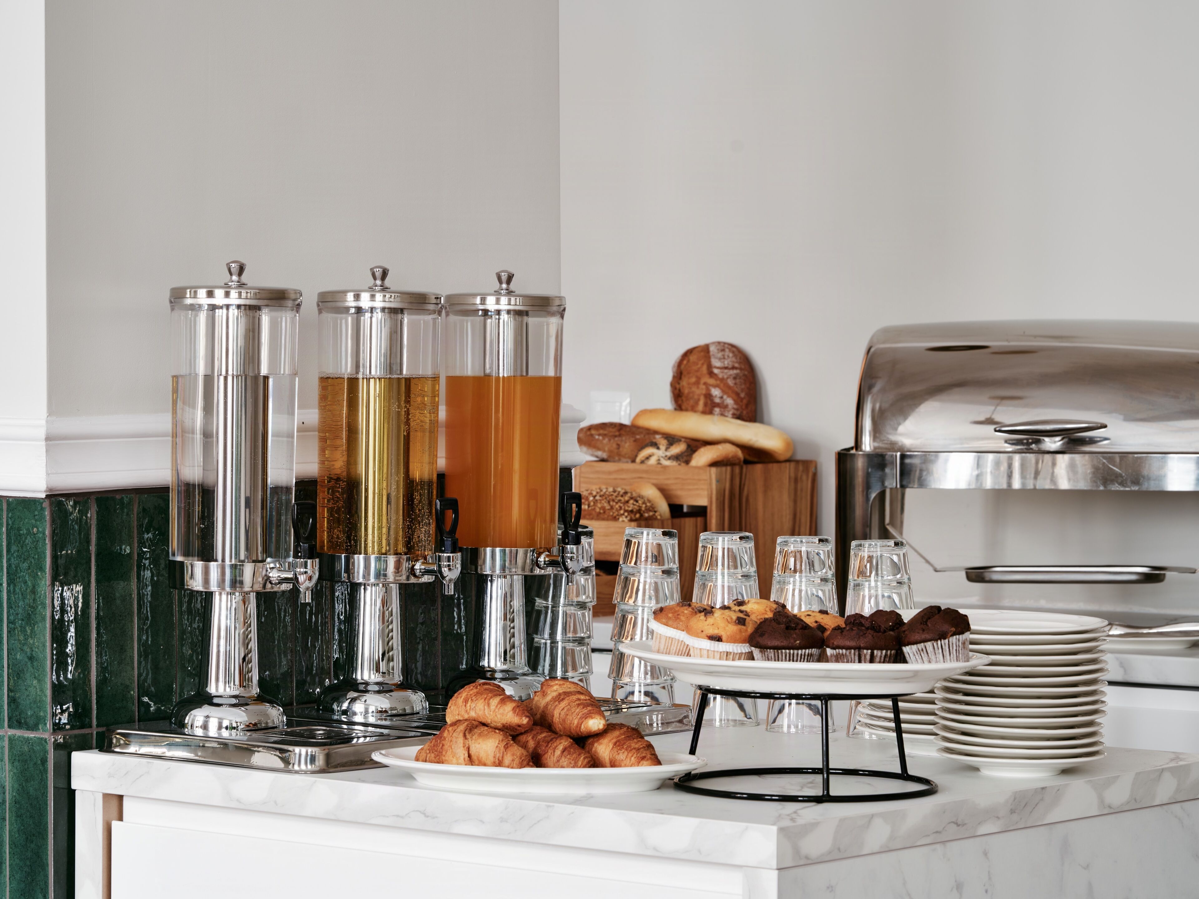 A breakfast buffet with coffee dispensers, pastries, and a bread display.