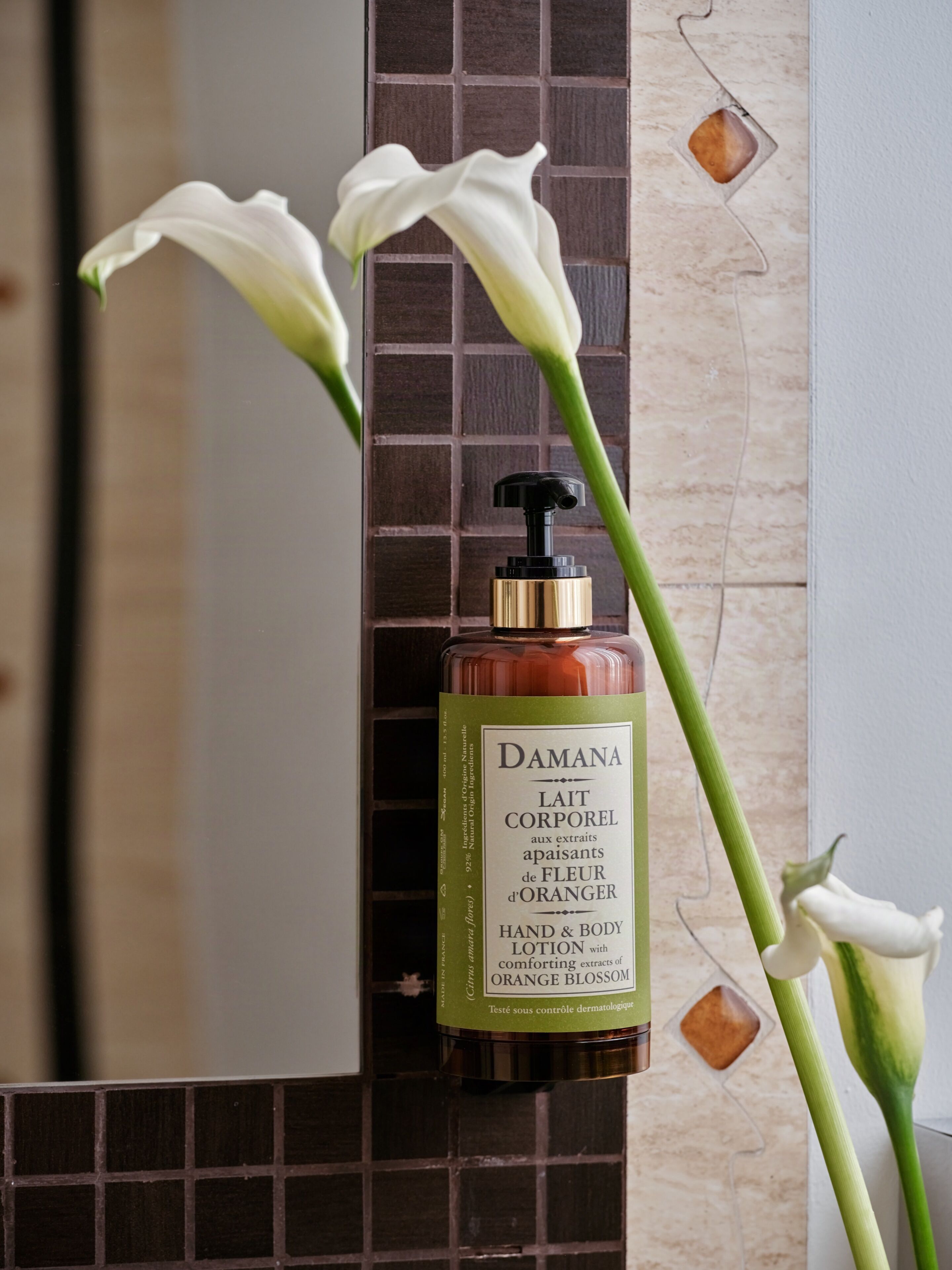 Eine Flasche Damana Hand- und Körperlotion mit Extrakt aus Orangenblüten auf einer gefliesten Oberfläche, zusammen mit einem Spiegel und weißen Blüten.