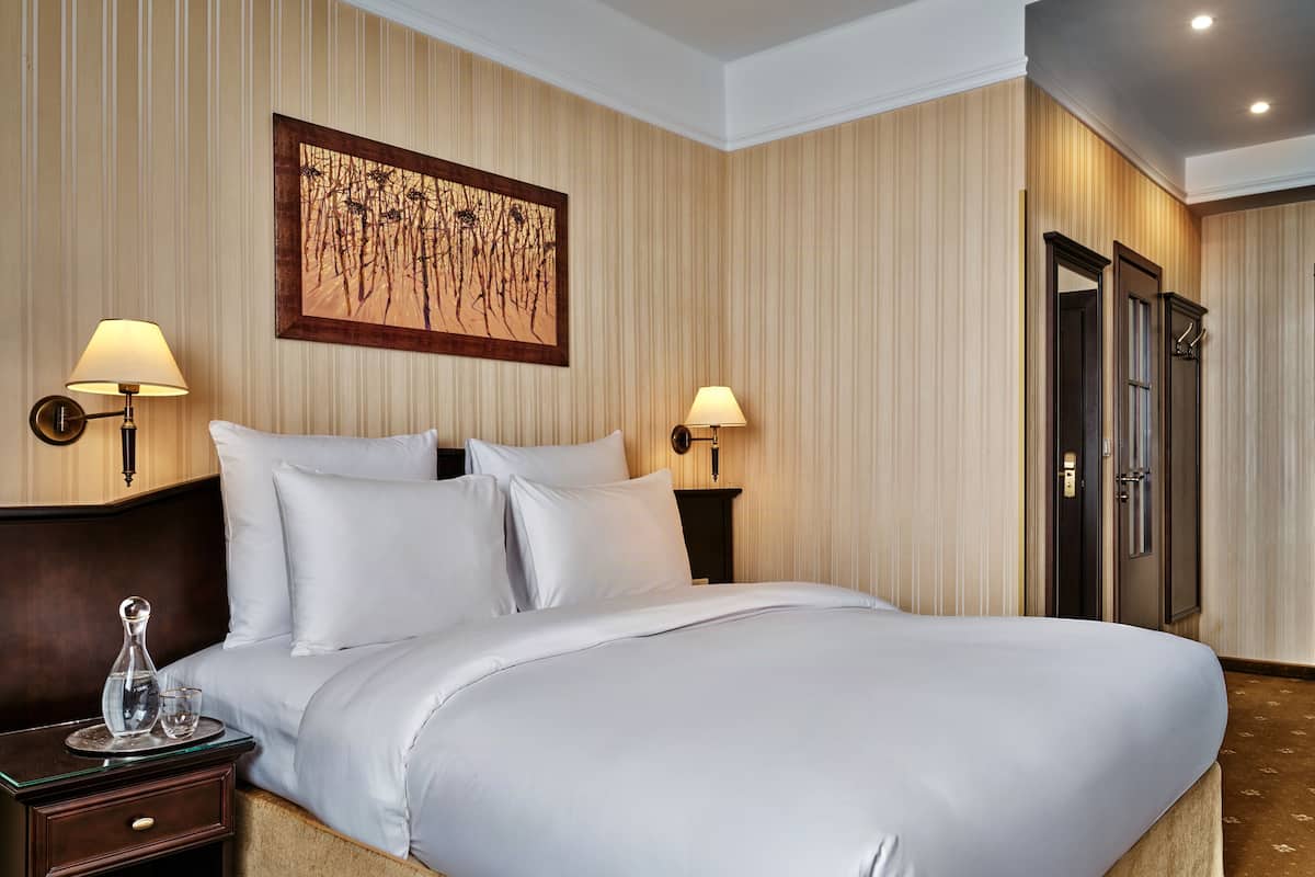 Una habitación de hotel con una cama grande, dos lámparas fijadas a la pared, una obra de arte enmarcada, una mesita de noche de madera oscura y una puerta con picaporte.