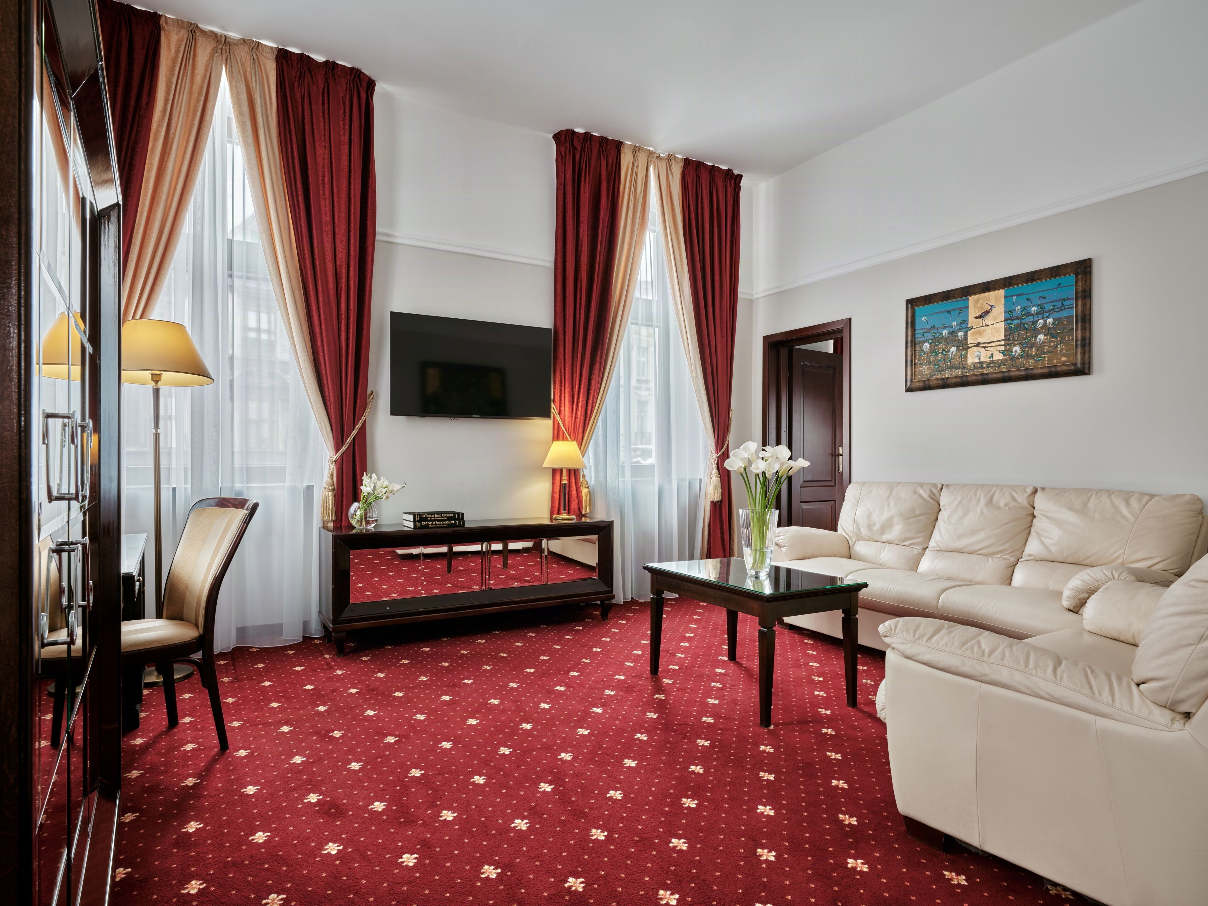 Una habitación de hotel con alfombra roja, un sofá, un televisor, una mesa con un florero y cortinas.