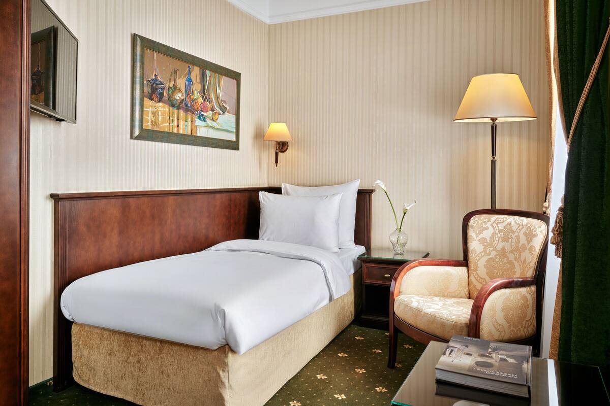 Una habitación de hotel con cama, una silla, una mesita de noche, una lámpara, un jarrón con flores y un cuadro en la pared.