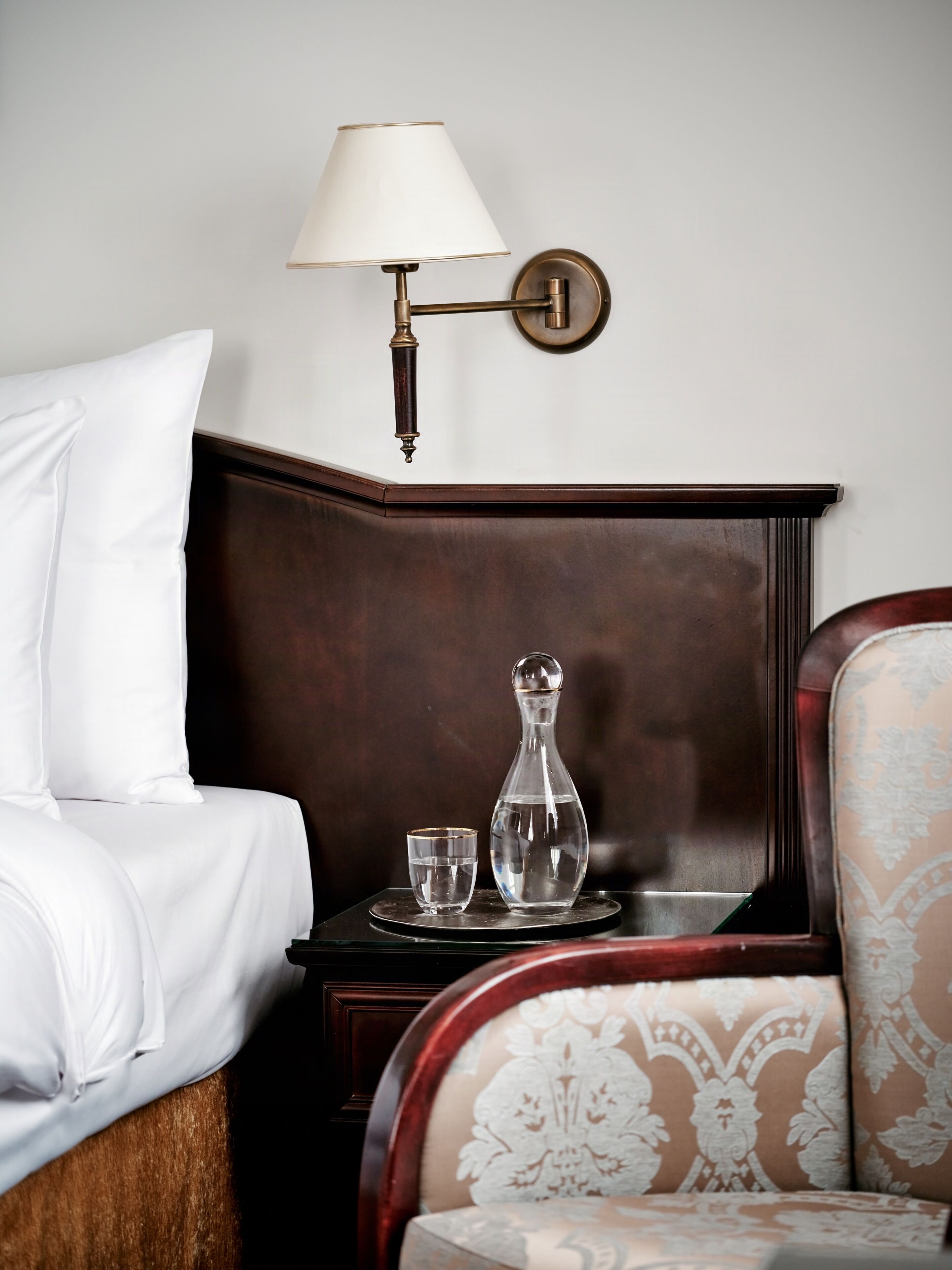 Una cama bien tendida con sábanas blancas, una lámpara de noche, una mesita con jarrón y taza de vidrio, y un sillón con estampado.