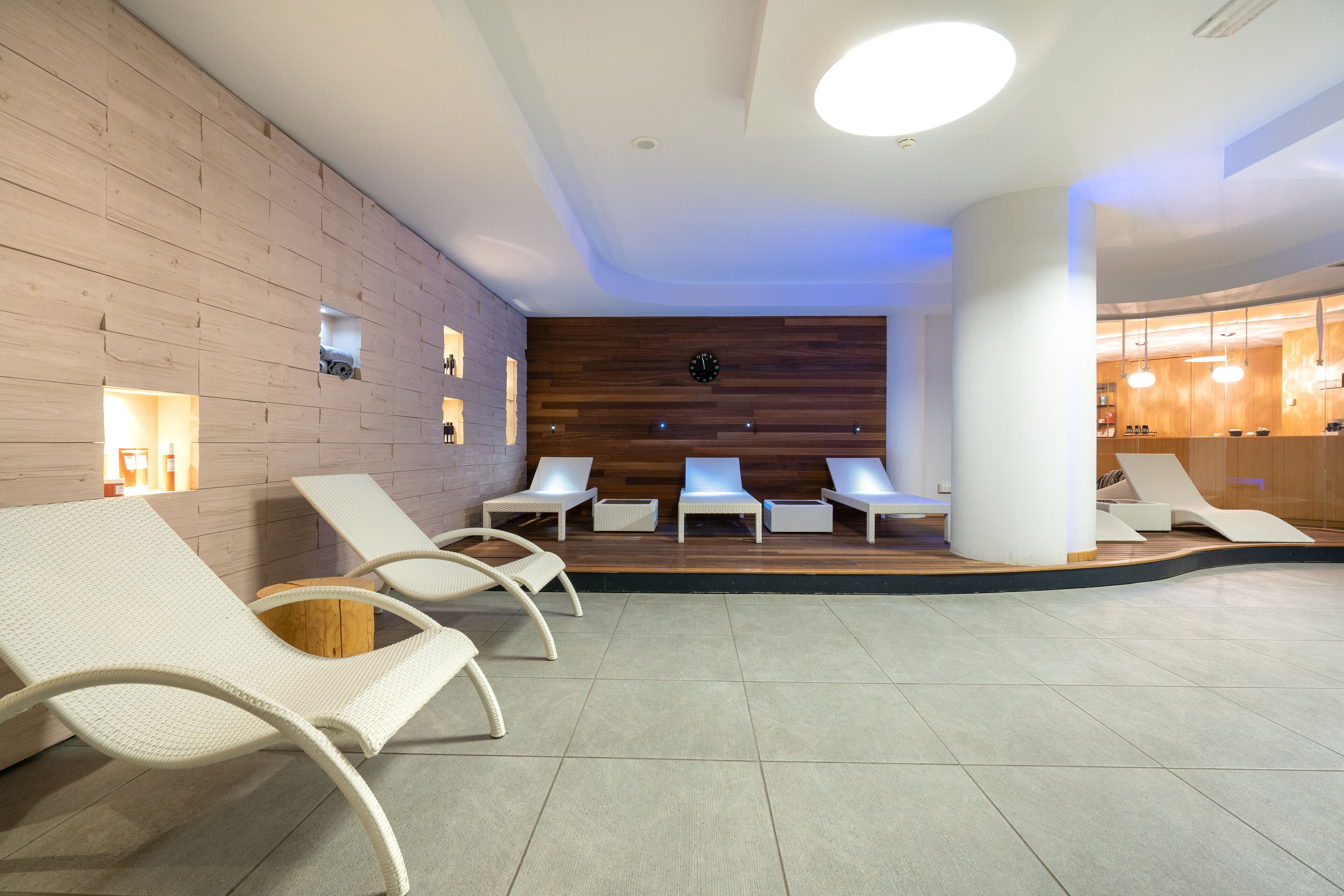 Un espace de détente intérieur moderne, avec des fauteuils blancs, un mur en bois doté d’étagères intégrées et une grande horloge.