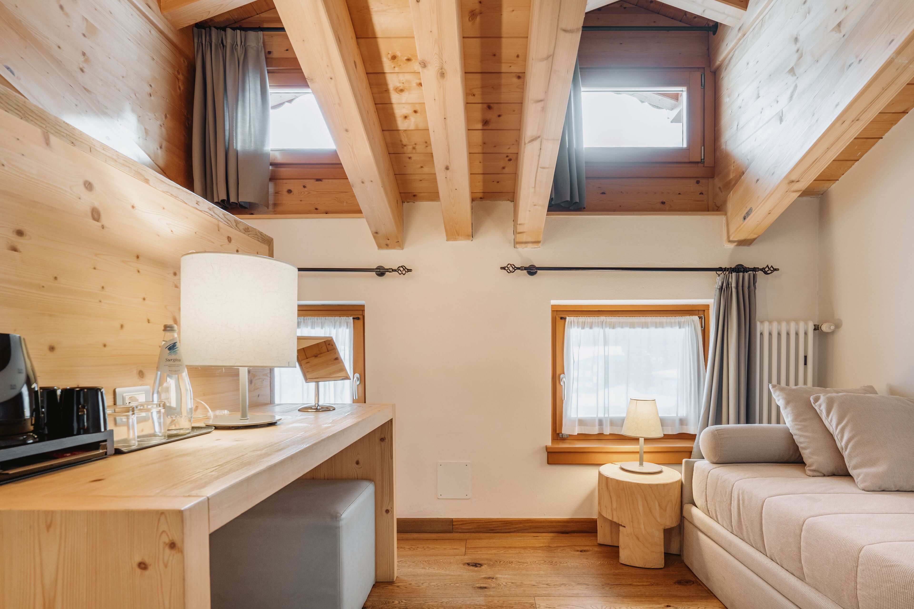 Una habitación acogedora con vigas de madera, un escritorio con lámpara, un sofá y una ventana con cortinas.