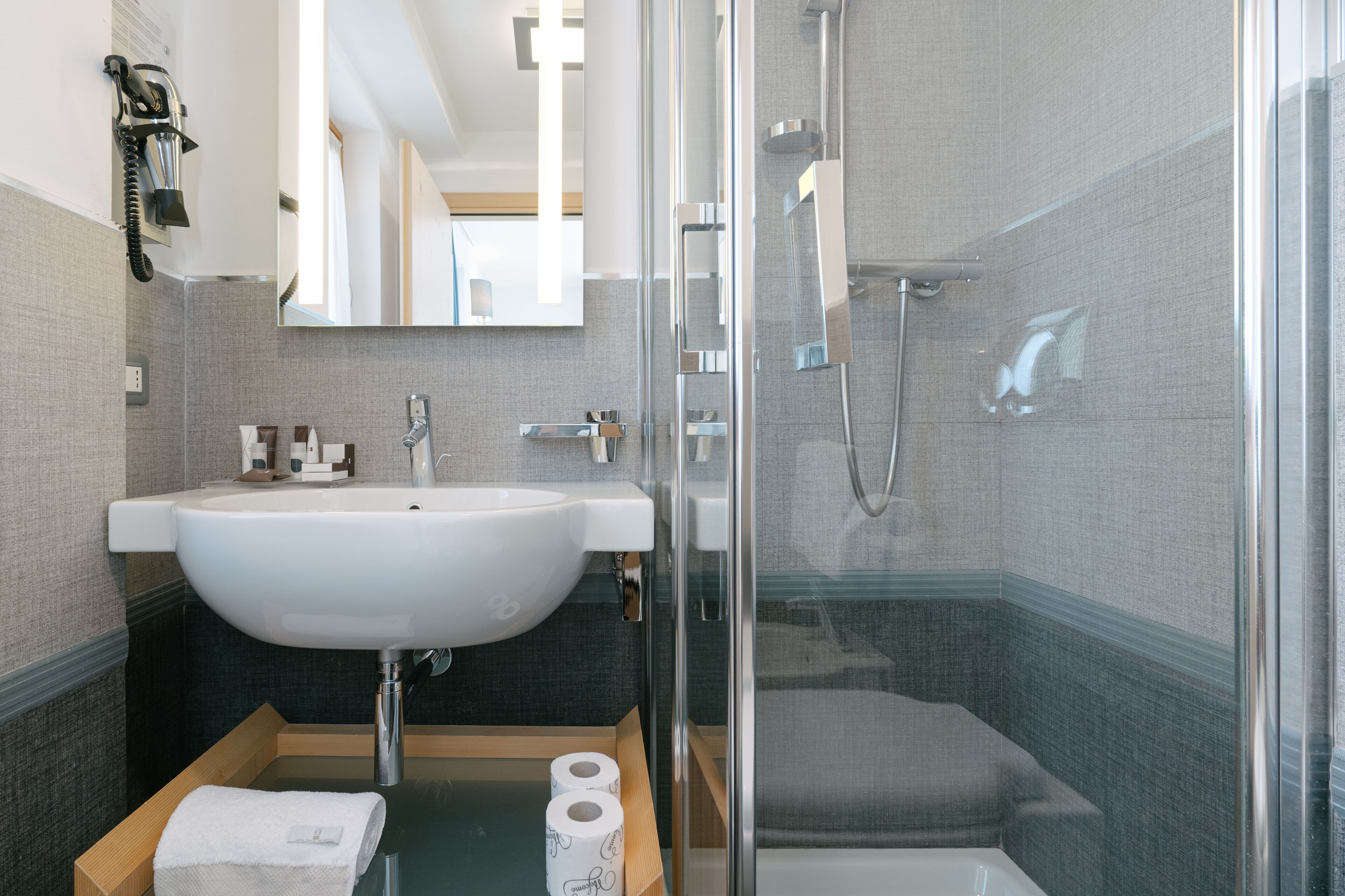 A modern bathroom with a large white sink, a glass shower enclosure, and a wooden vanity with a towel and toilet paper.