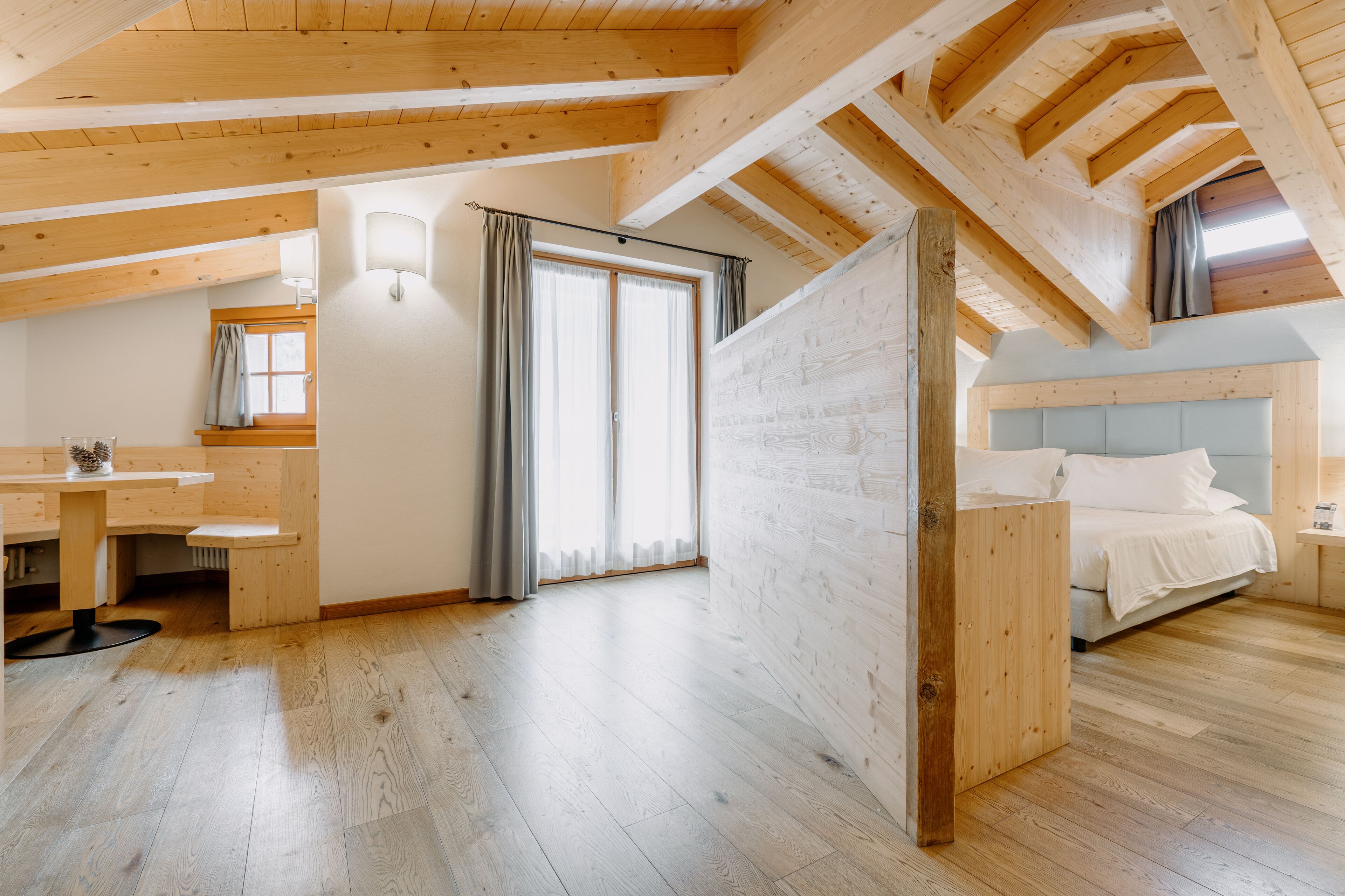 Un amplio dormitorio en el ático con vigas de madera, un ventanal con cortinas, una cama y un pequeño escritorio.