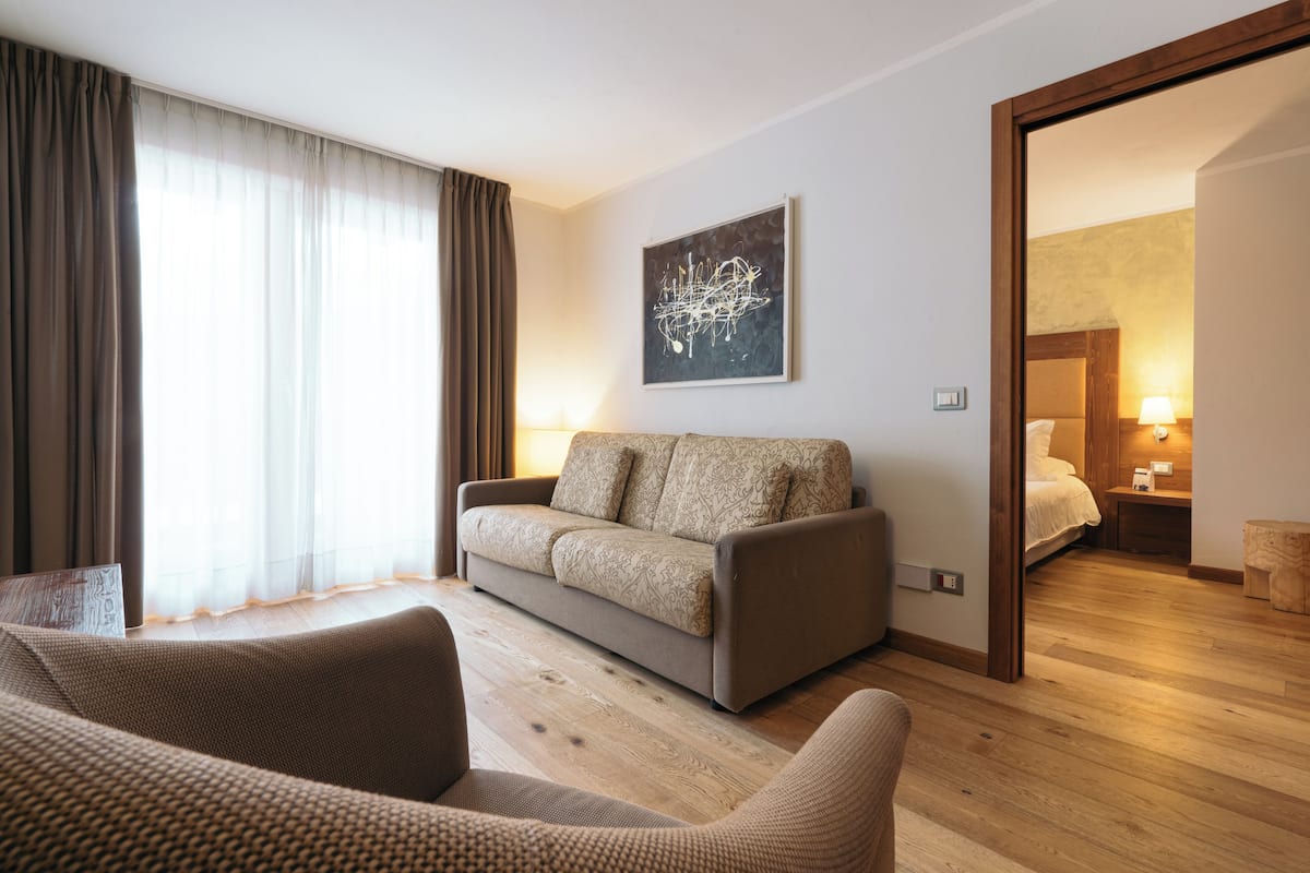 A modern hotel room with a sofa, a wooden floor, a painting on the wall, and a view into another room.