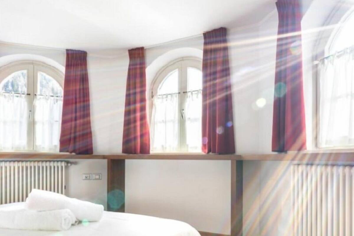 A hotel room with a bed, windows with curtains, a radiator, and a wooden floor.