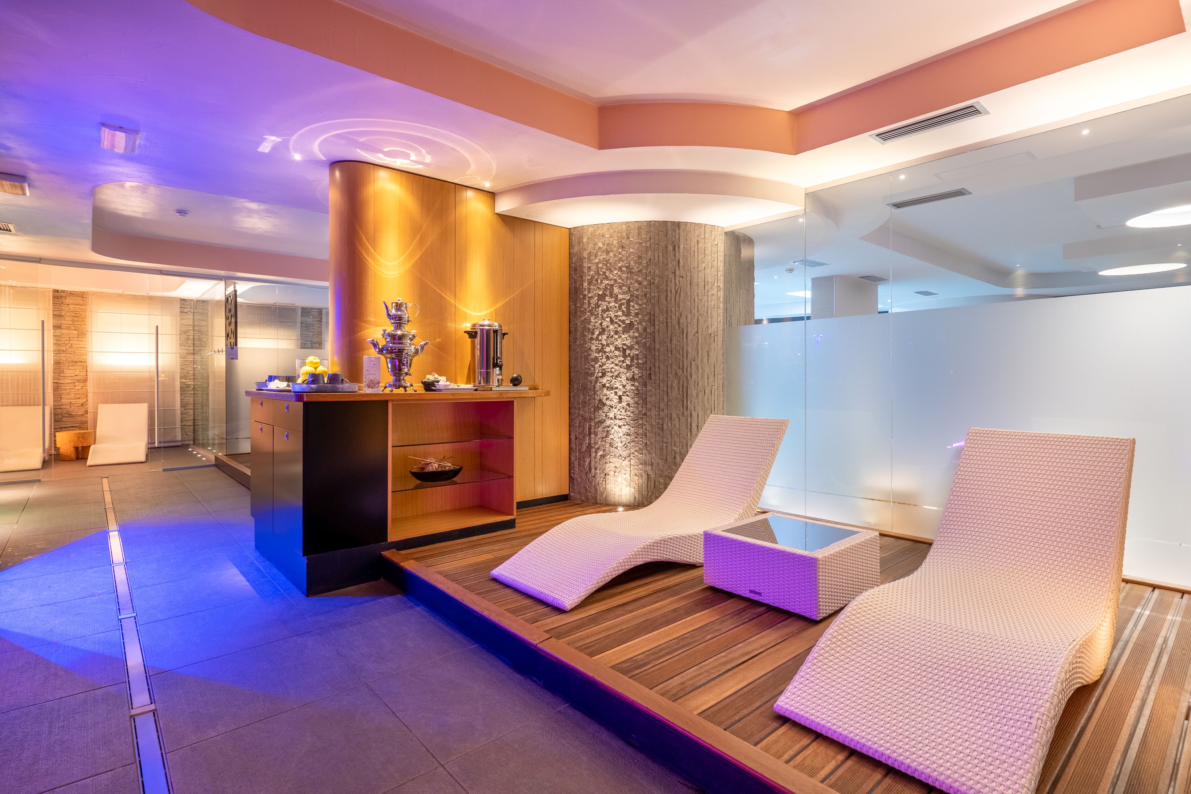 A modern spa area with wooden flooring, two white cushioned chairs, a wooden cabinet with a decorative item, and a textured wall.