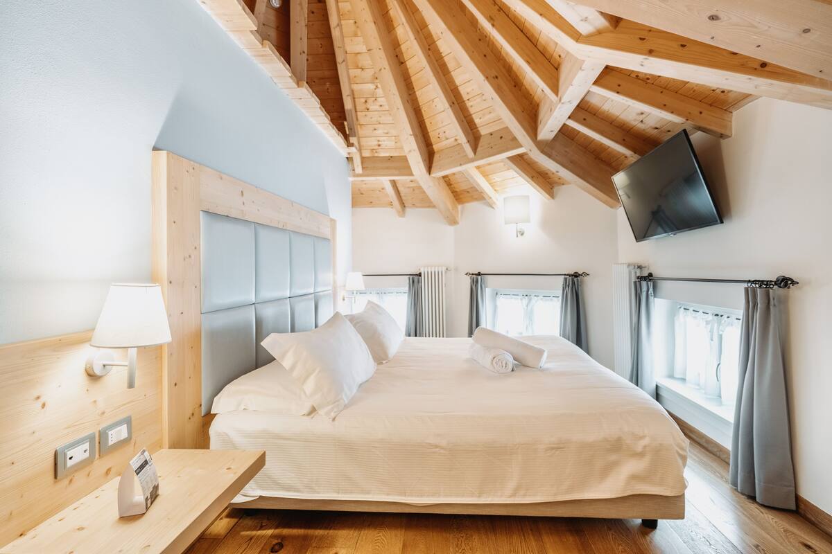 A bedroom with a wooden ceiling, a bed with white linens, a bedside table with a lamp, and a TV mounted on the wall.