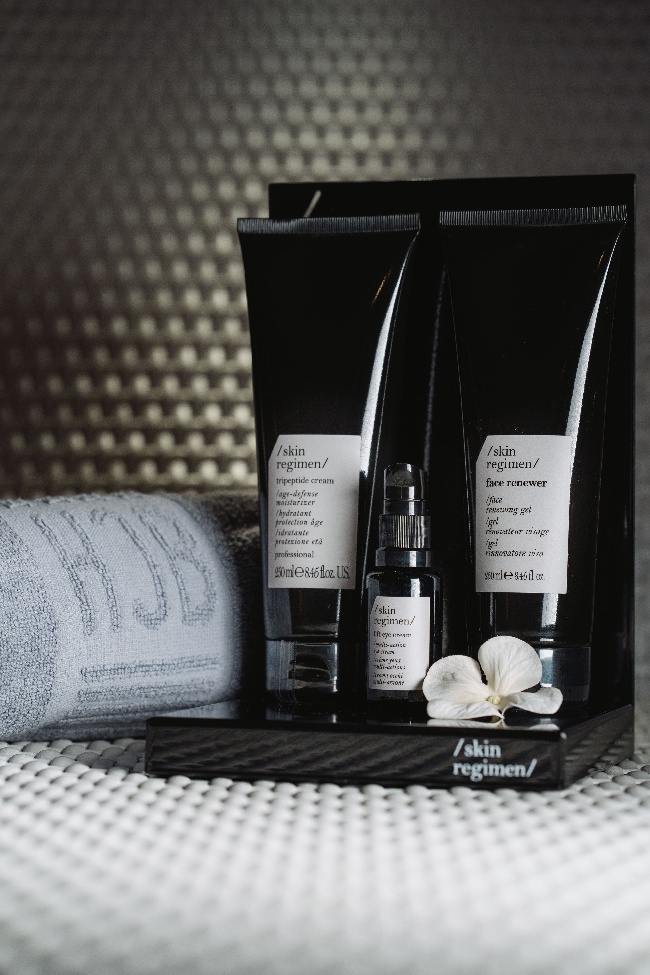 Un set de cuidado facial con tres productos sobre una bandeja, incluyendo una crema para el rostro y un sérum, colocado sobre una superficie texturizada con una flor blanca.
