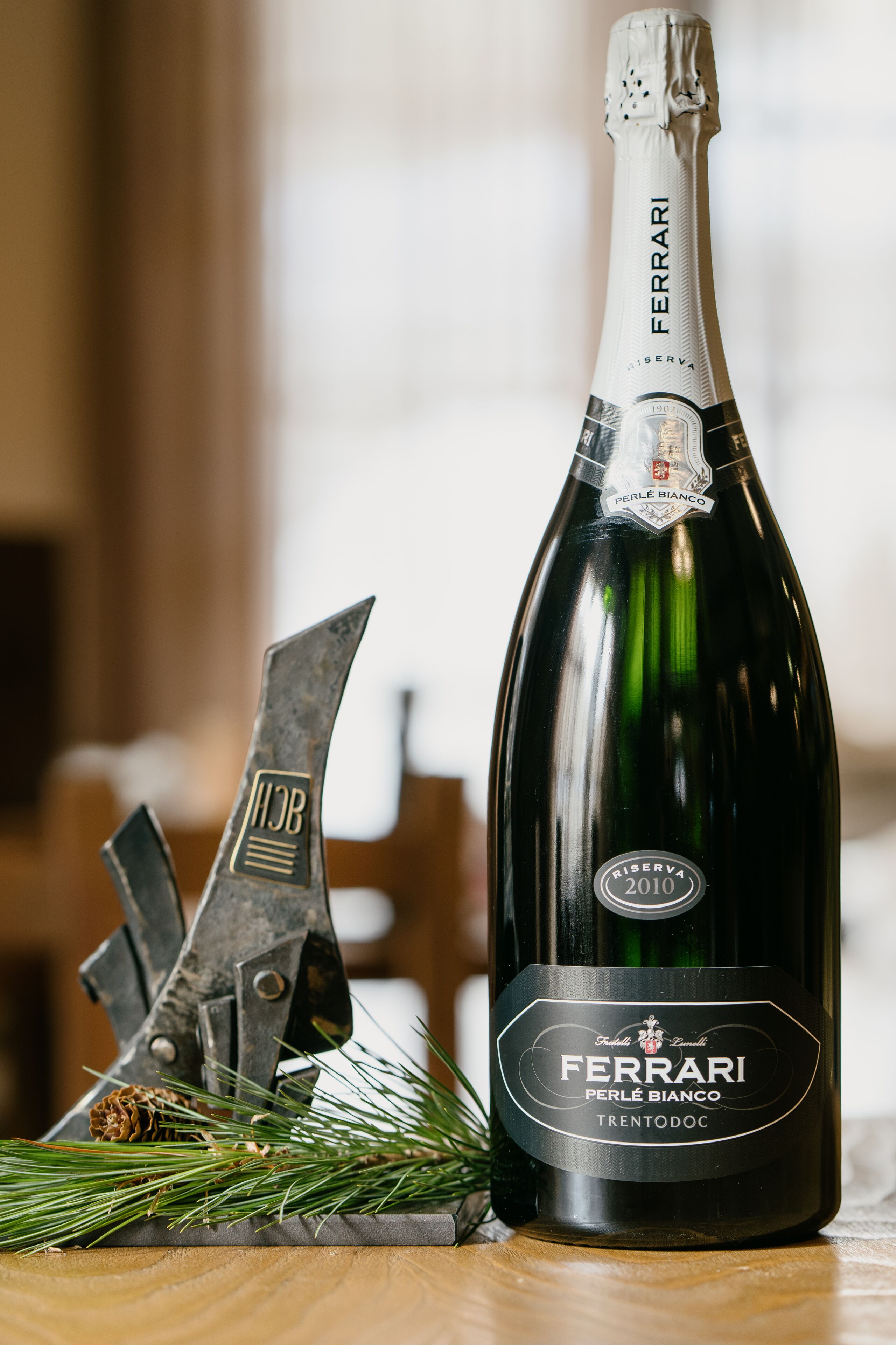 A bottle of Ferrari Perle Bianco wine with a label indicating it is a 2010 vintage, placed on a wooden surface with pine branches and a decorative metal sculpture.